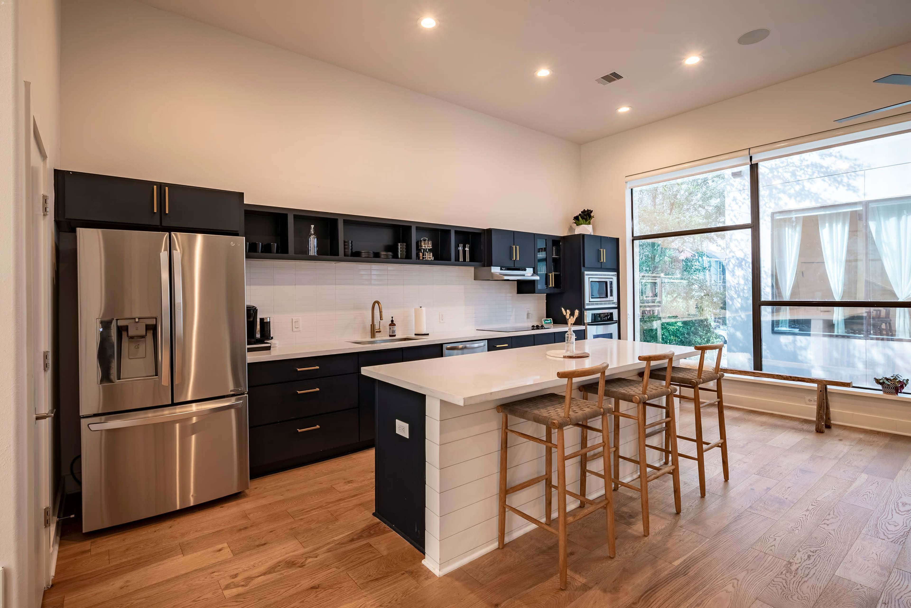 The image shows a modern kitchen with dark cabinetry, stainless steel appliances, a large island with bar stools, and a large window providing natural light.