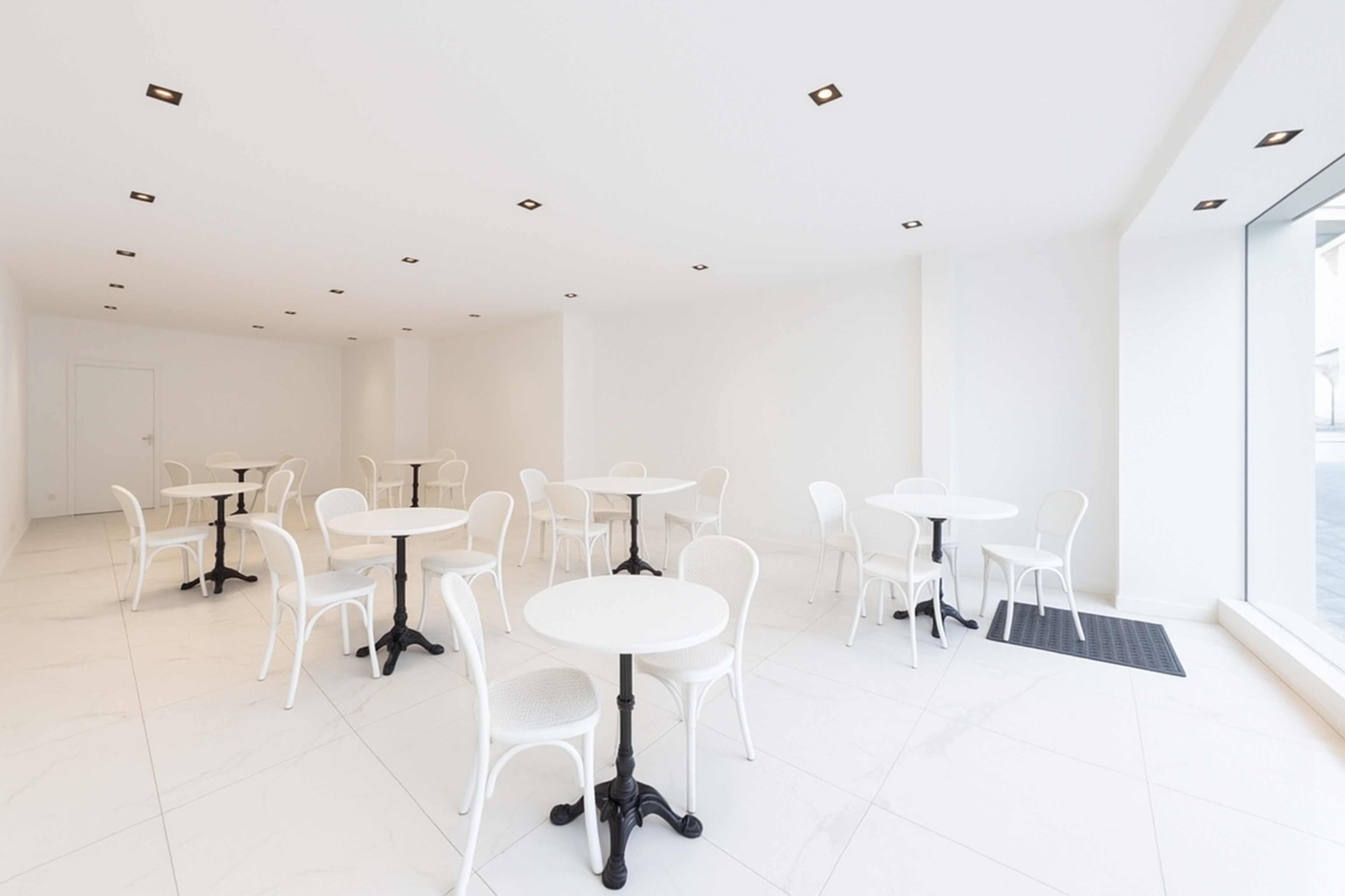 The image shows a spacious, minimalistic cafe with multiple white round tables and chairs arranged on a light-colored tiled floor.