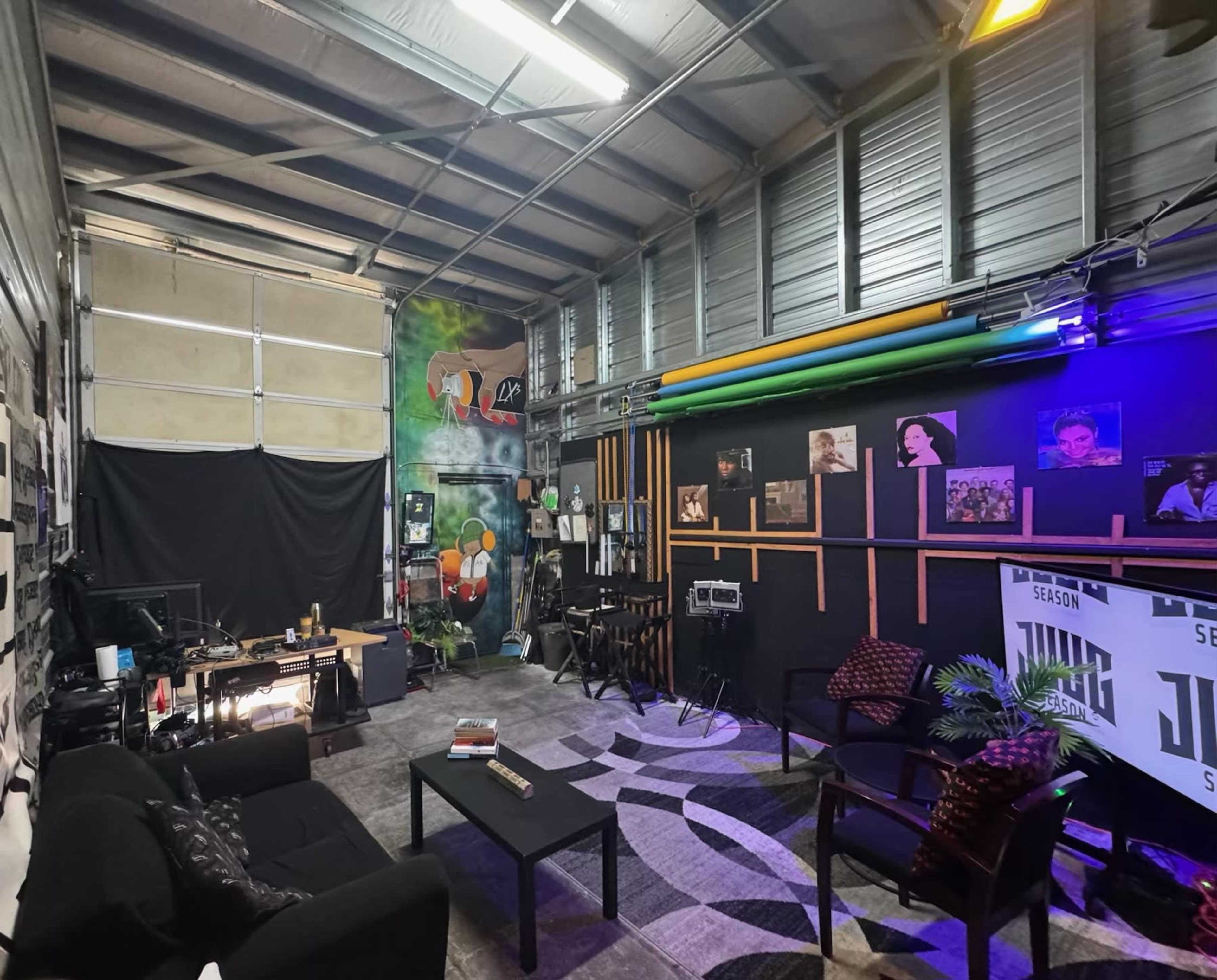 The image shows an indoor studio space featuring a seating area with a black couch, chairs, and a coffee table, along with colorful wall art and various photographs displayed on the walls.