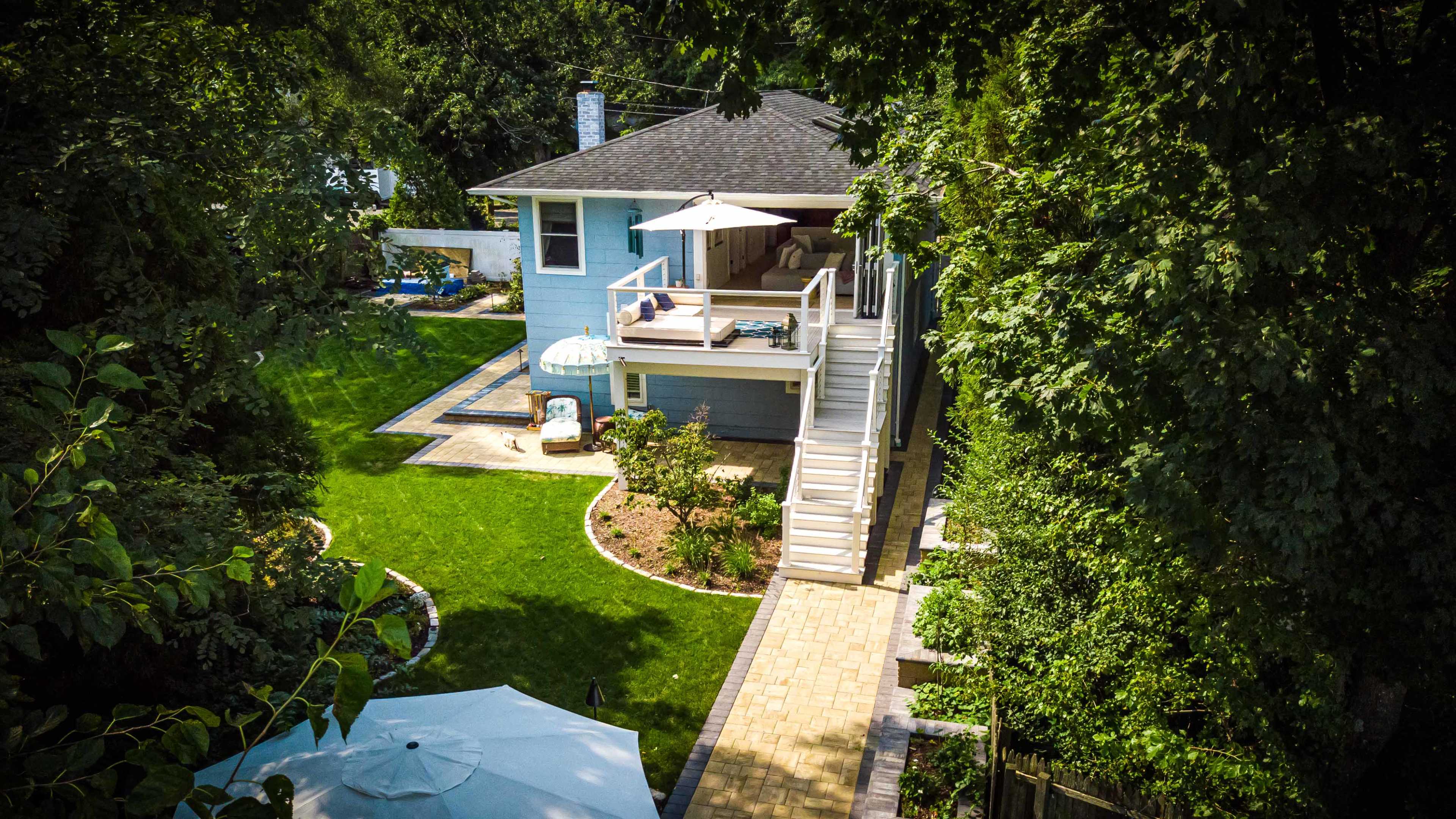 The image shows a two-story blue house with a spacious backyard, featuring a patio, a pathway, and landscaped greenery.