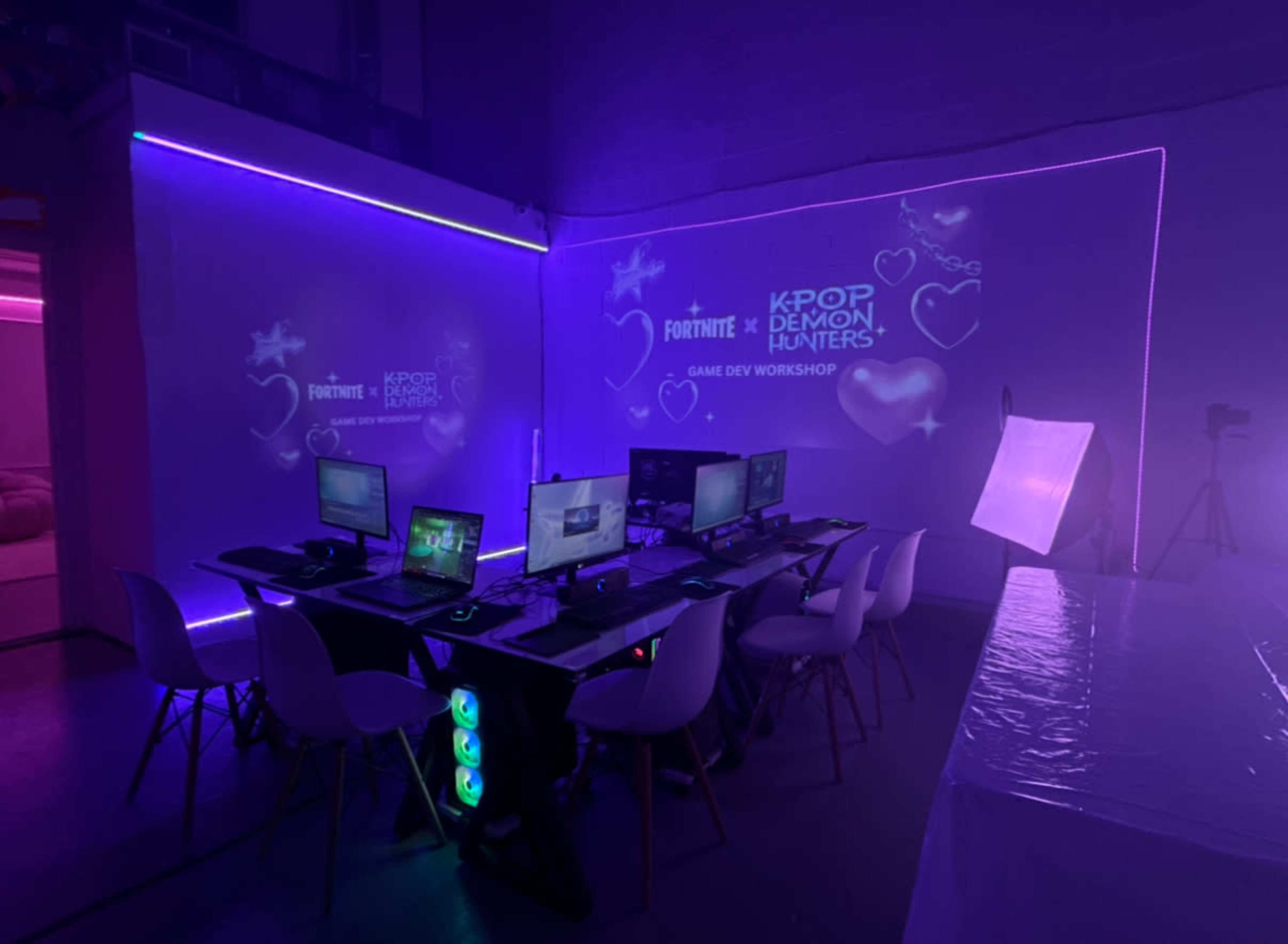 The image shows a dimly lit workspace with several computer desks arranged in a semi-circle, featuring glowing purple lights and a projection of "KPOP DEMON HUNTERS GAME DEV WORKSHOP" on the wall.