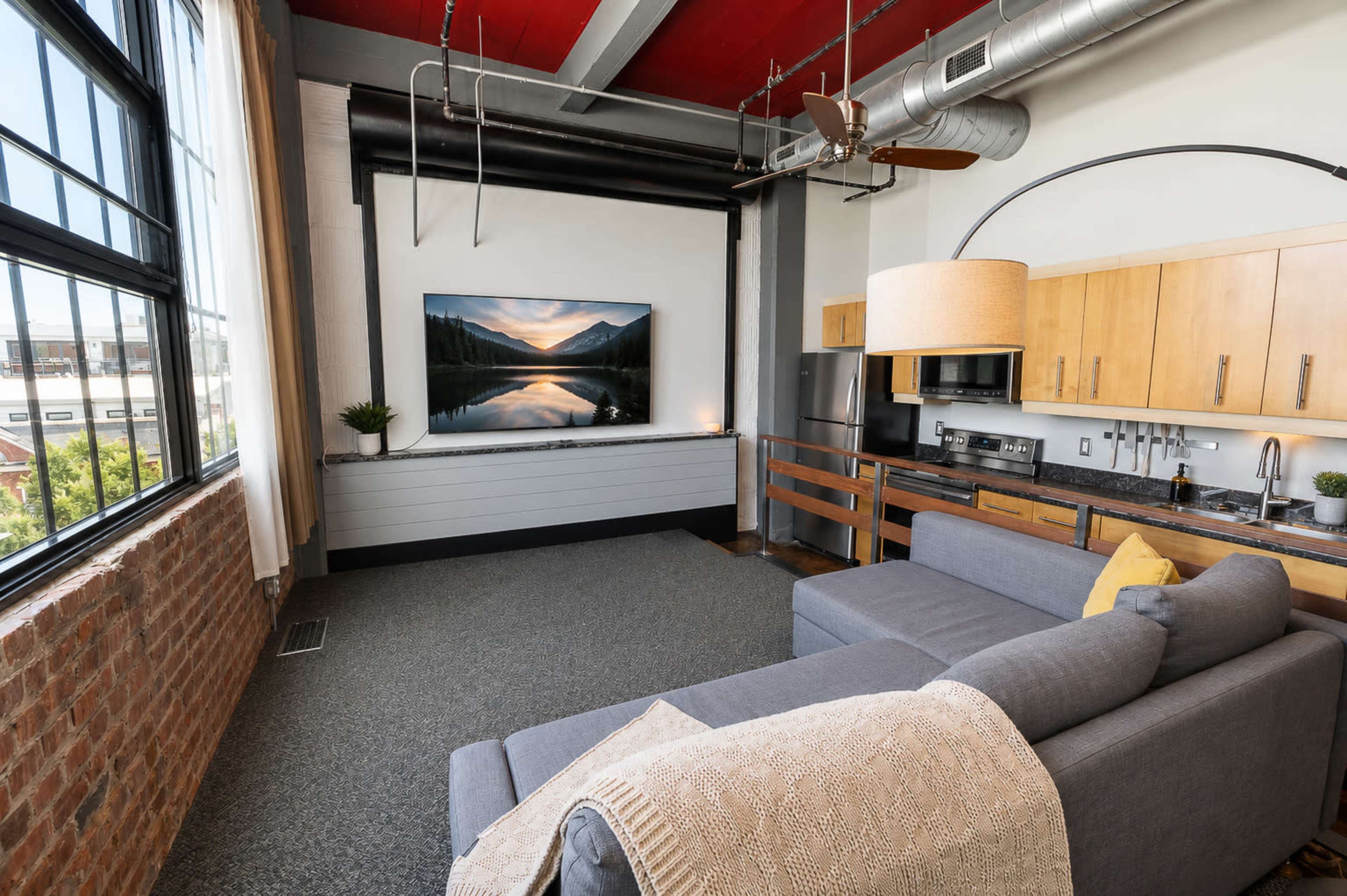 The image shows a modern loft-style living area with a gray sectional sofa, a large wall-mounted television, and wooden kitchen cabinetry against a backdrop of exposed brick walls and high ceilings.