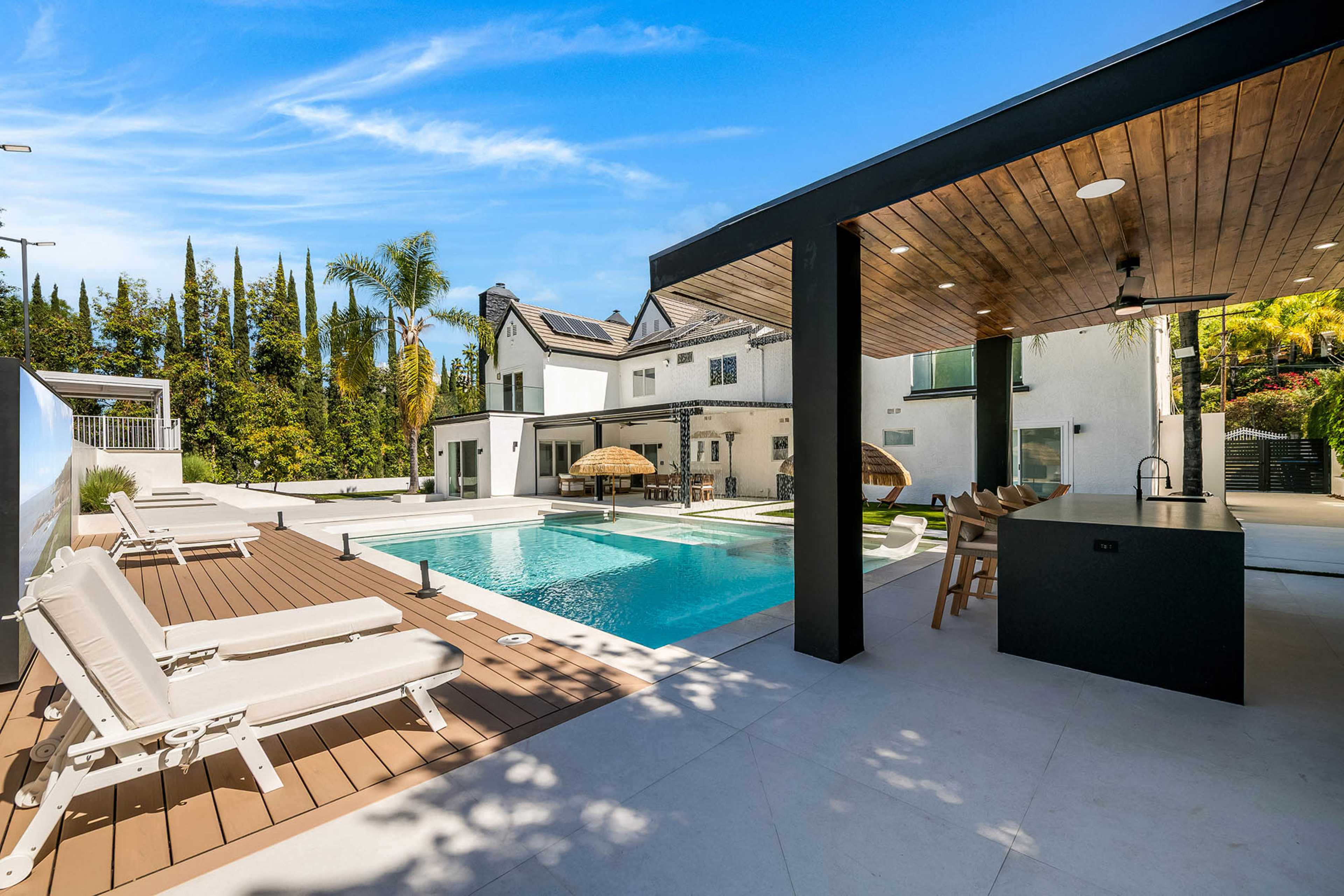 The image shows a modern outdoor space featuring a swimming pool, lounge chairs, and a covered seating area with a bar.
