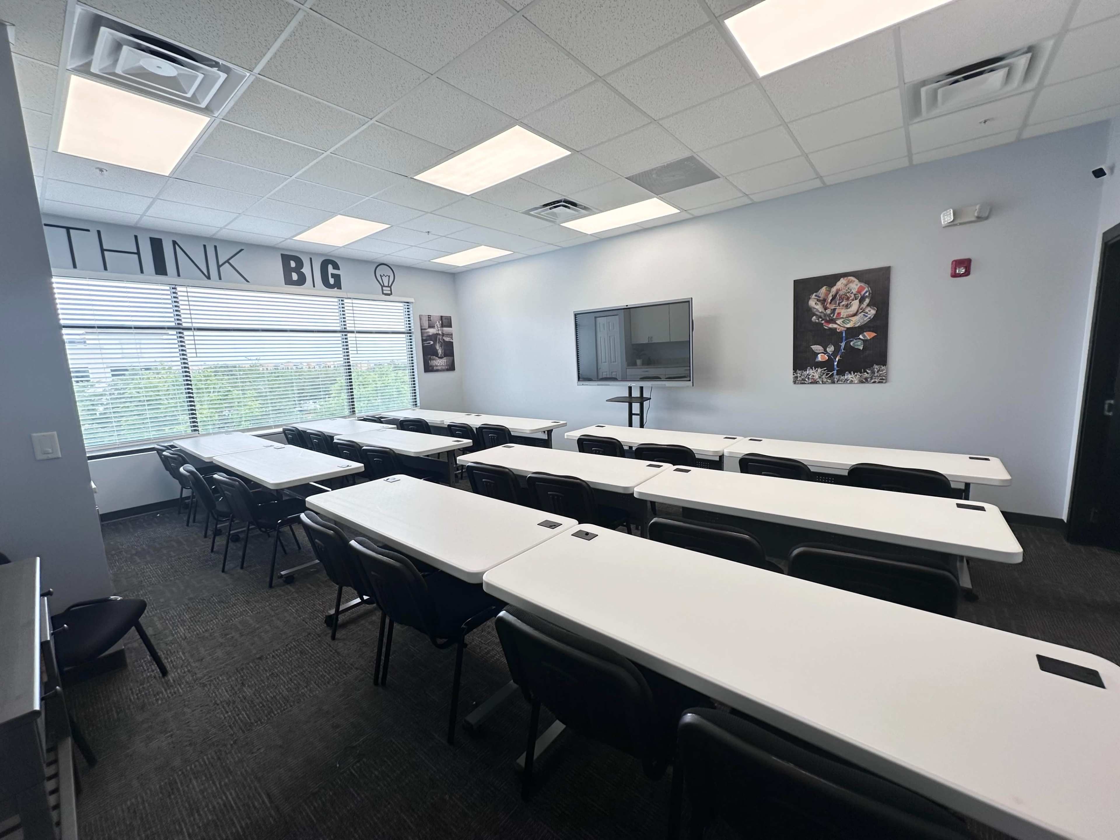 A modern classroom is set up with multiple tables and chairs arranged in rows, featuring large windows and a wall with motivational text.