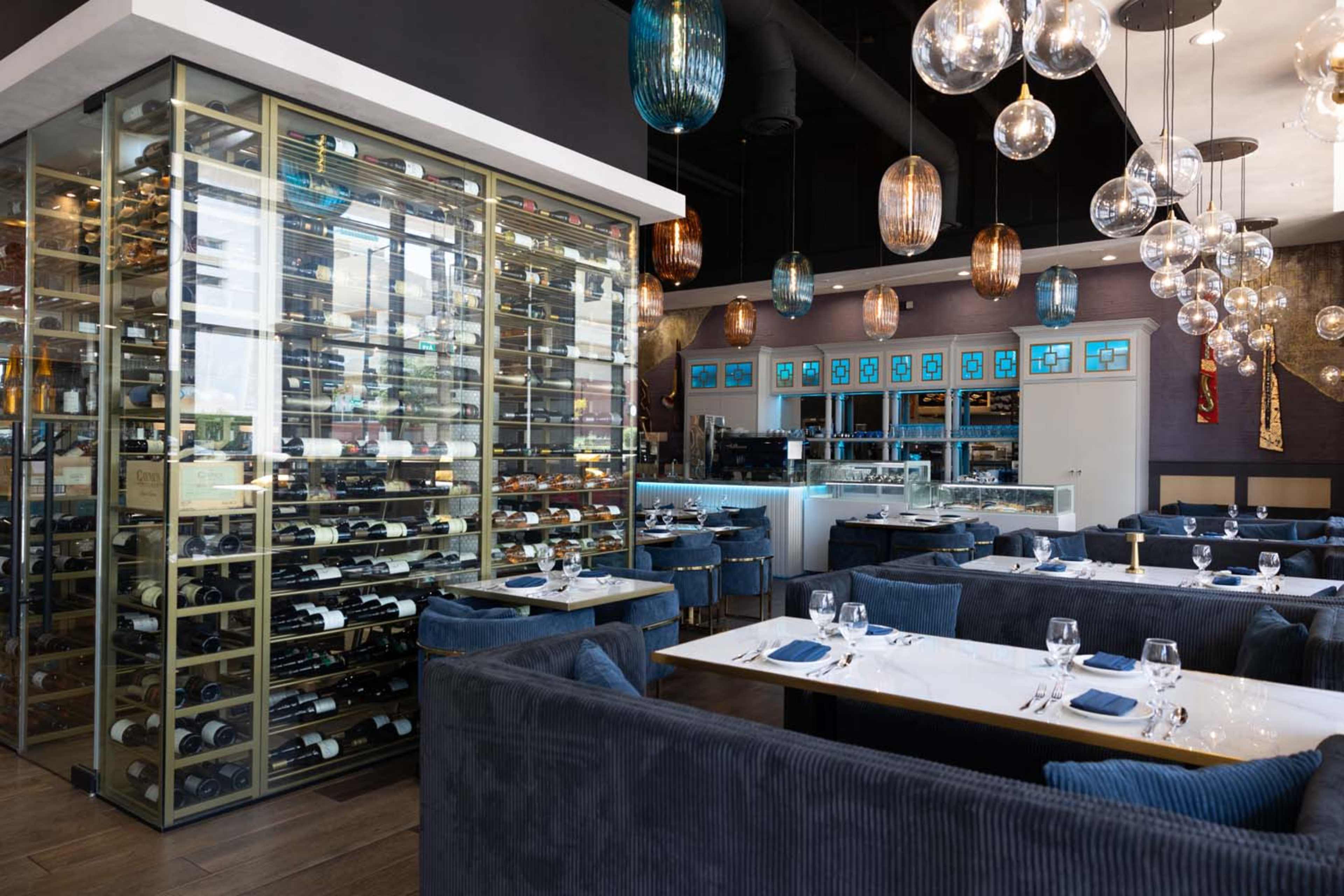 The image shows a modern restaurant interior featuring plush seating, a glass wine storage display, and decorative pendant lights hanging from the ceiling.