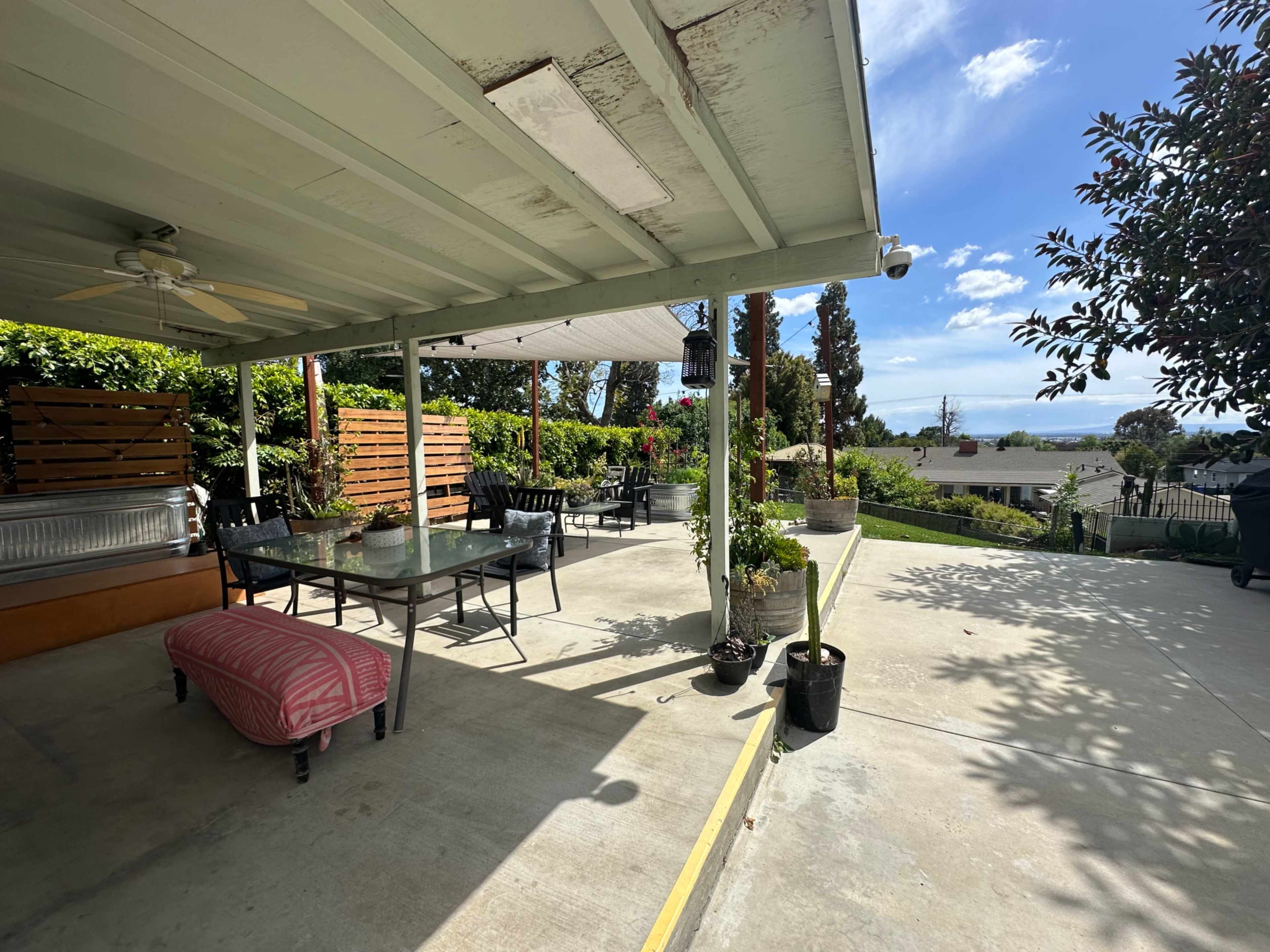 A patio area with a table and chairs, potted plants, and a view of a hillside under a partially cloudy sky.