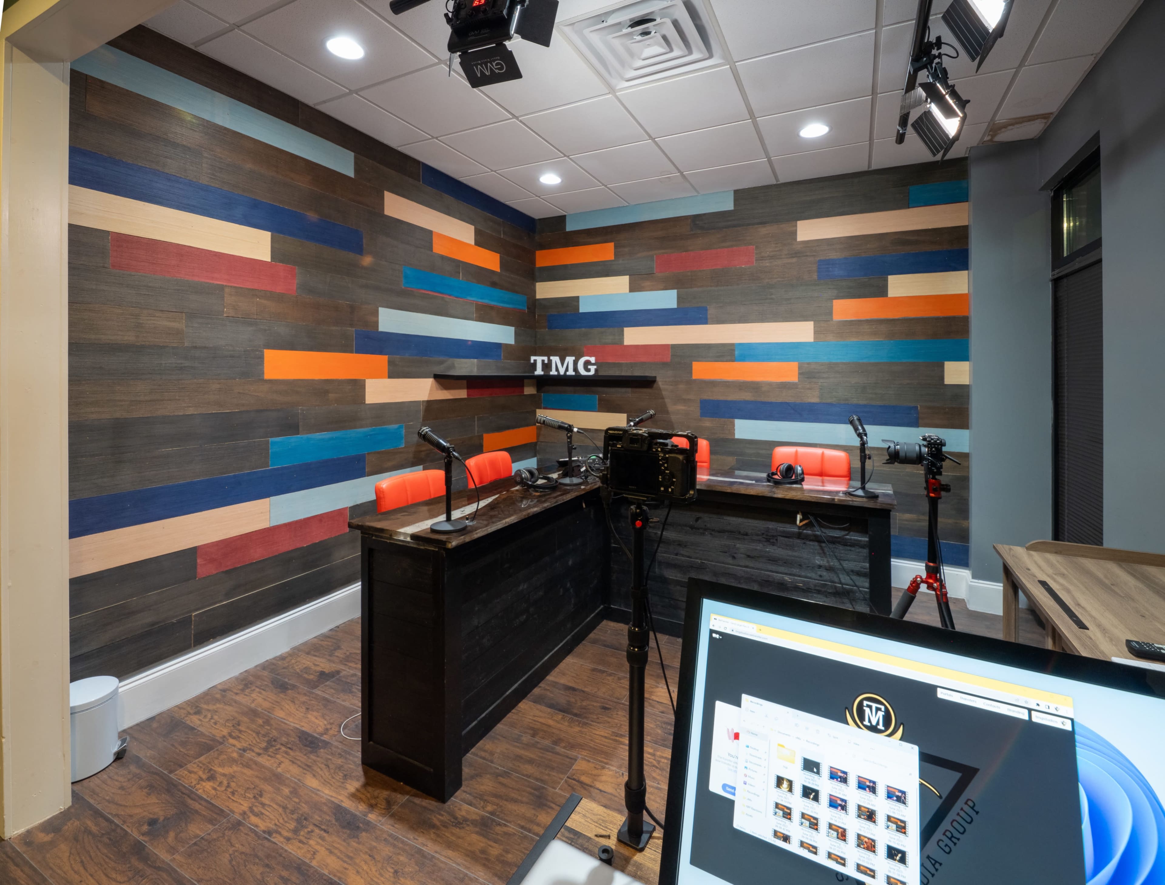 A modern recording studio with a colorful wooden wall, two orange chairs at a black table, and camera equipment set up for video recording.