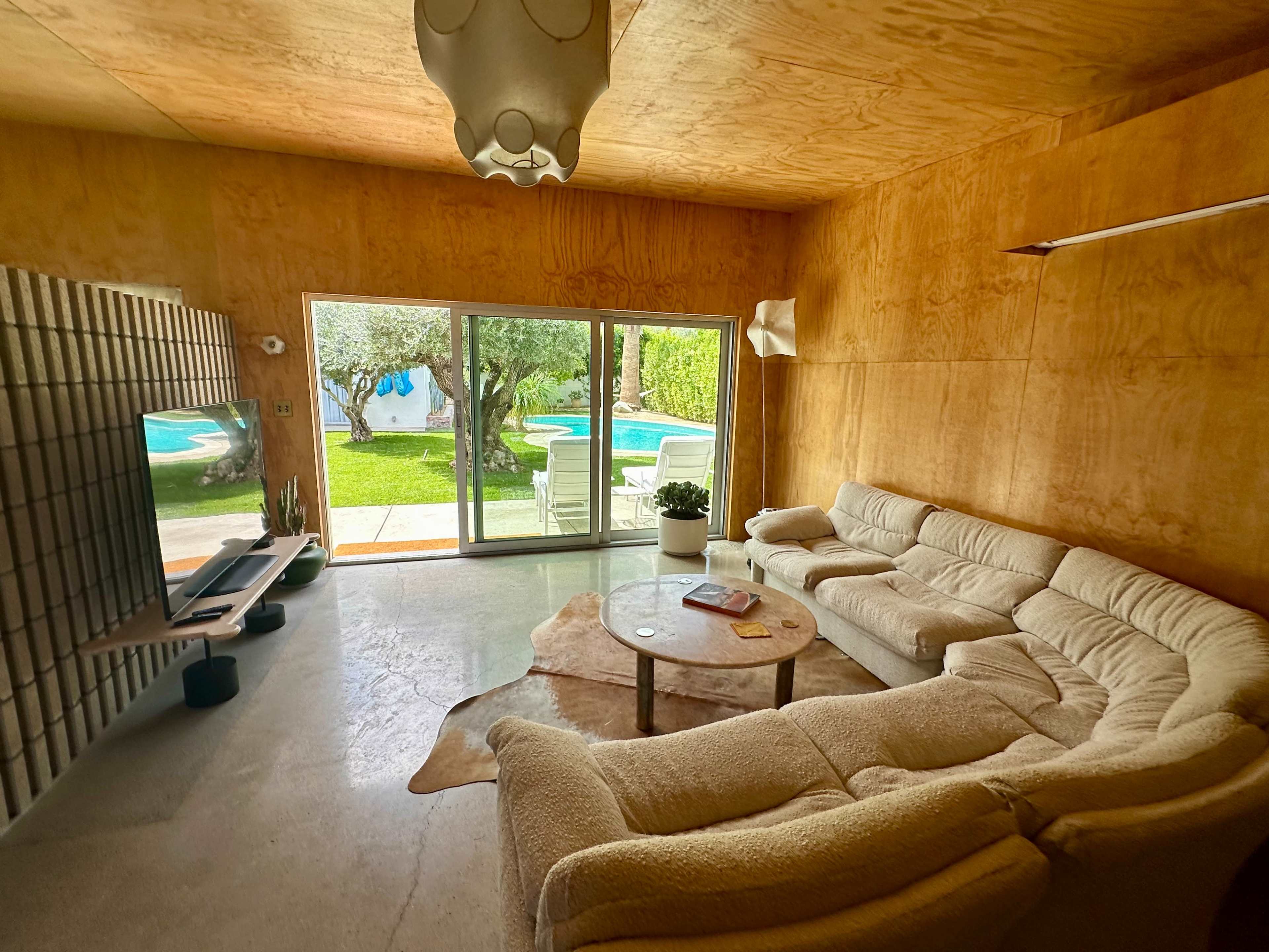 A cozy living room with a large curved sofa, a coffee table, and a view of a pool and garden through large glass doors.