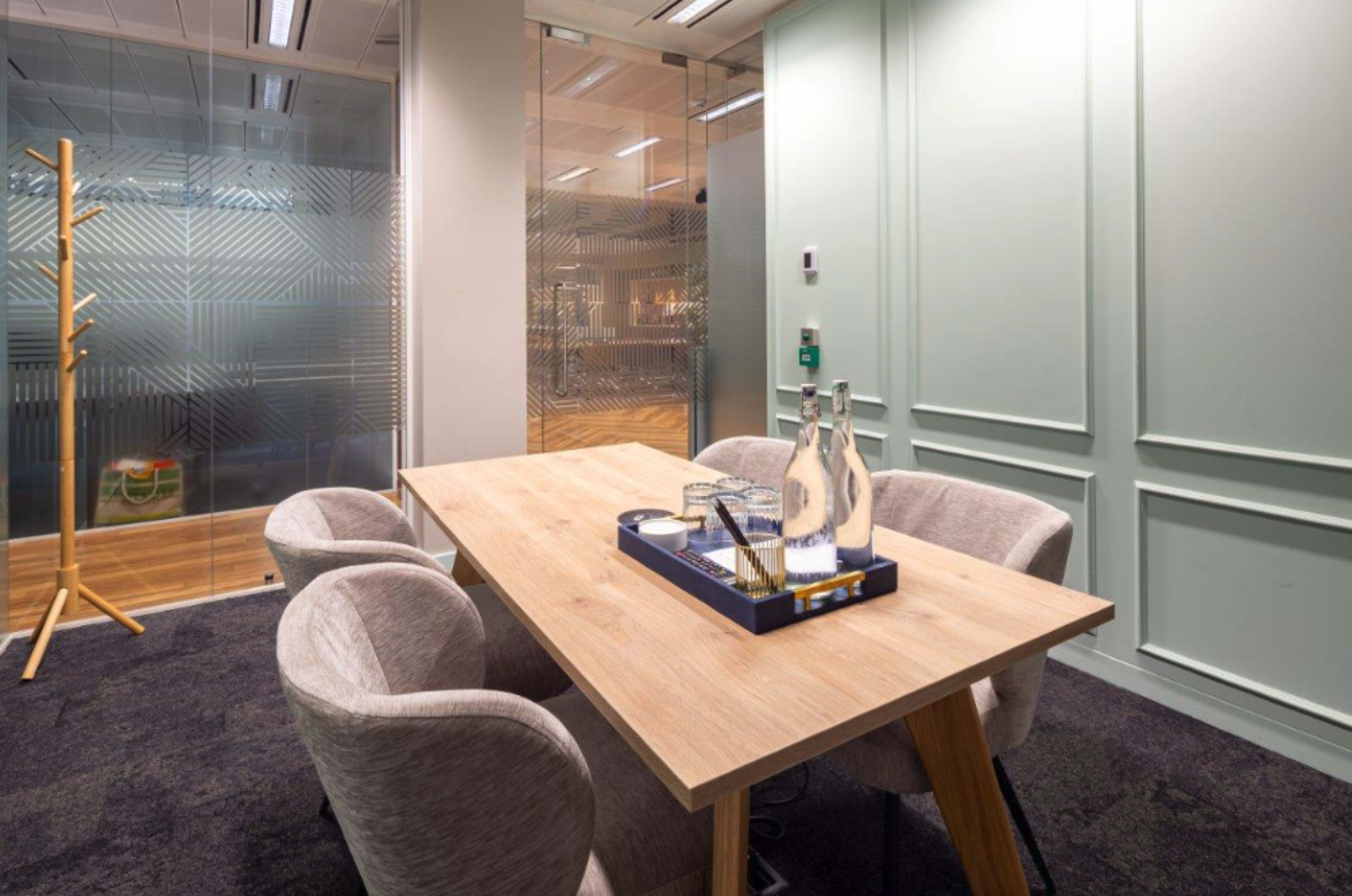 A wooden conference table is set with water bottles and glasses in a small, modern meeting room featuring pale green walls and contemporary furnishings.