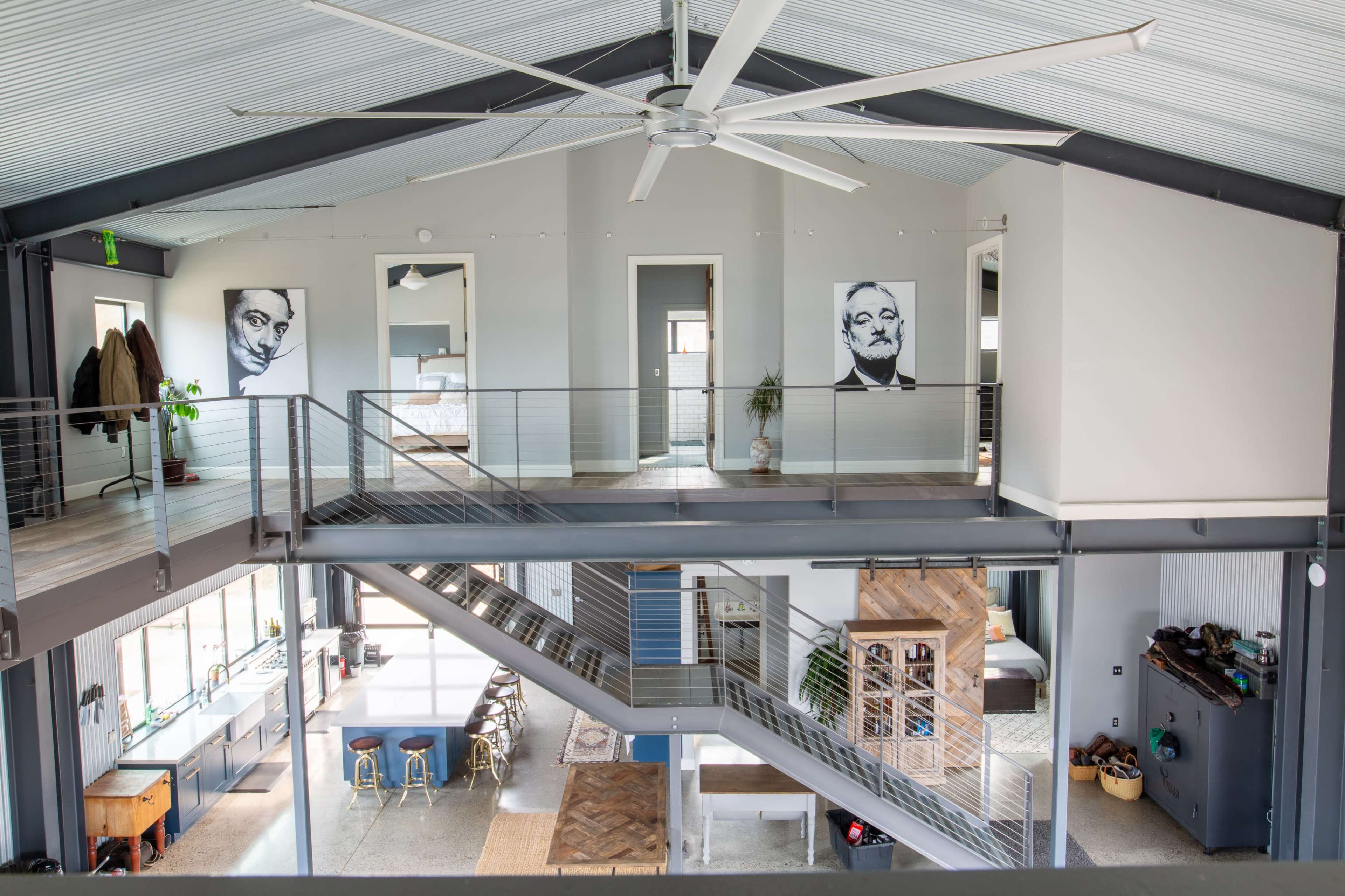 The image shows a modern open-concept interior with a central staircase, large ceiling fan, and framed portraits hanging on the walls.
