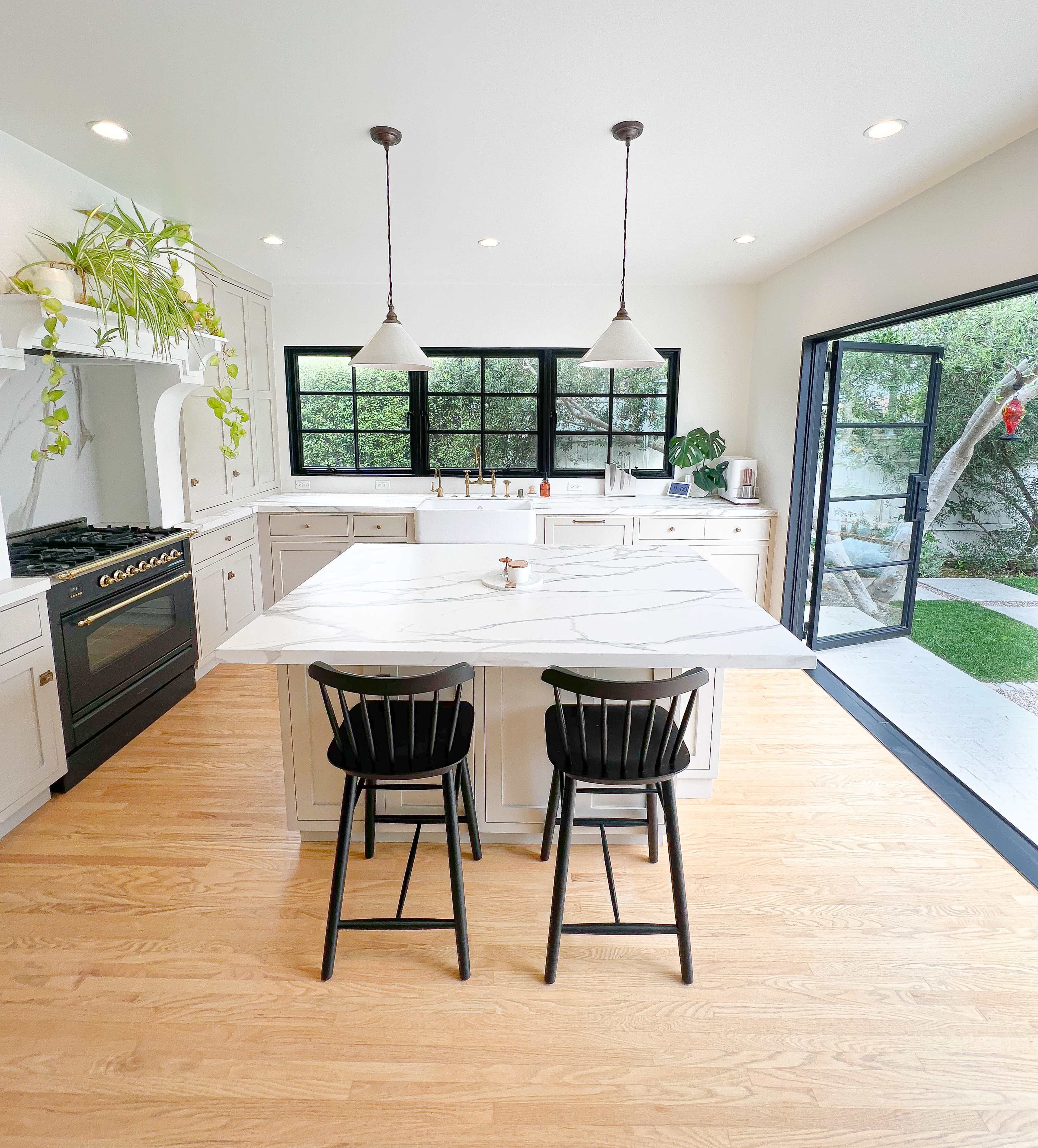A modern kitchen featuring a white marble island with two black stools, a gas stove, and large windows overlooking an outdoor space.