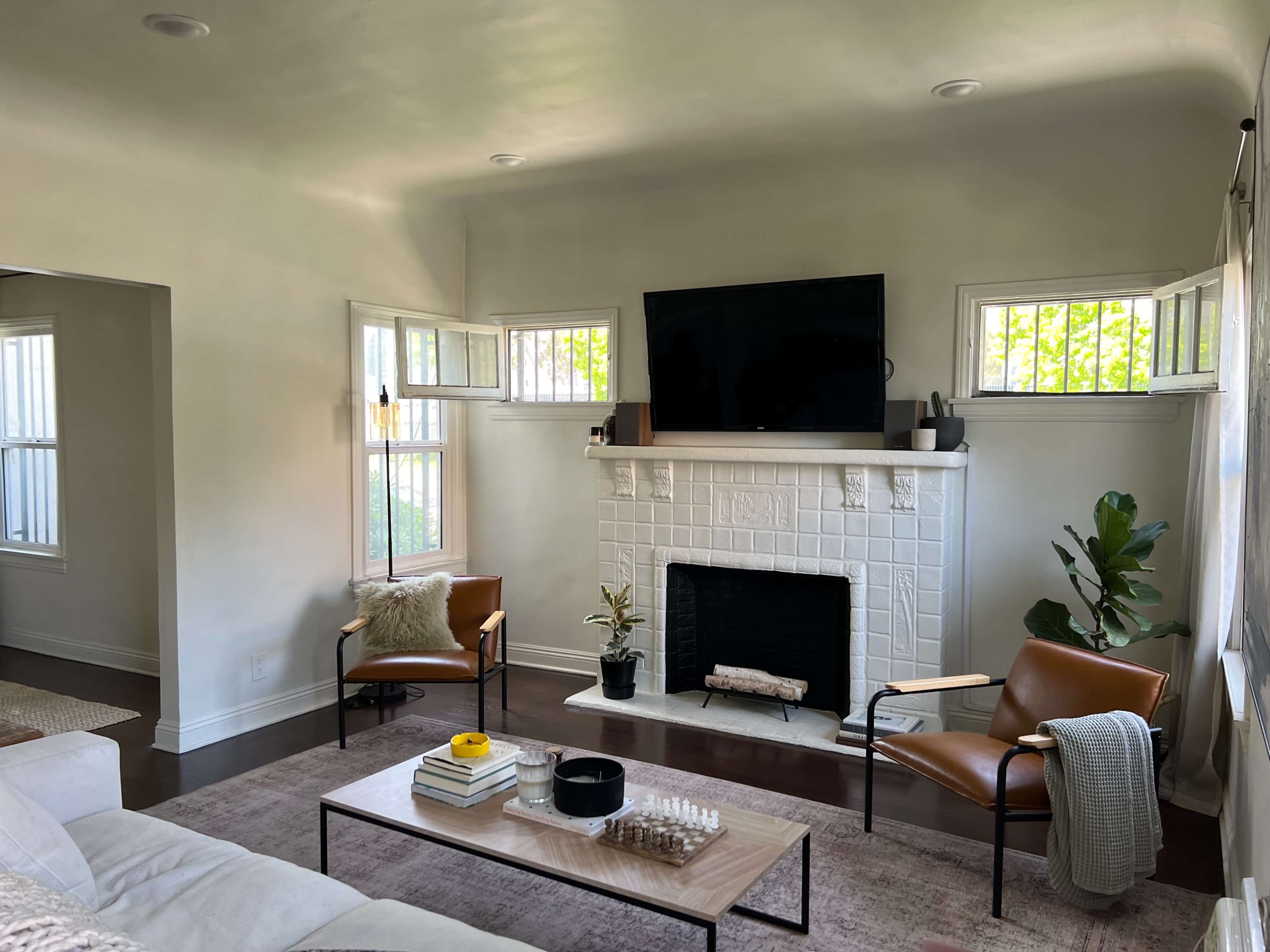 The image shows a bright living room featuring a white fireplace with a mounted television above it, accompanied by two chairs and a coffee table.