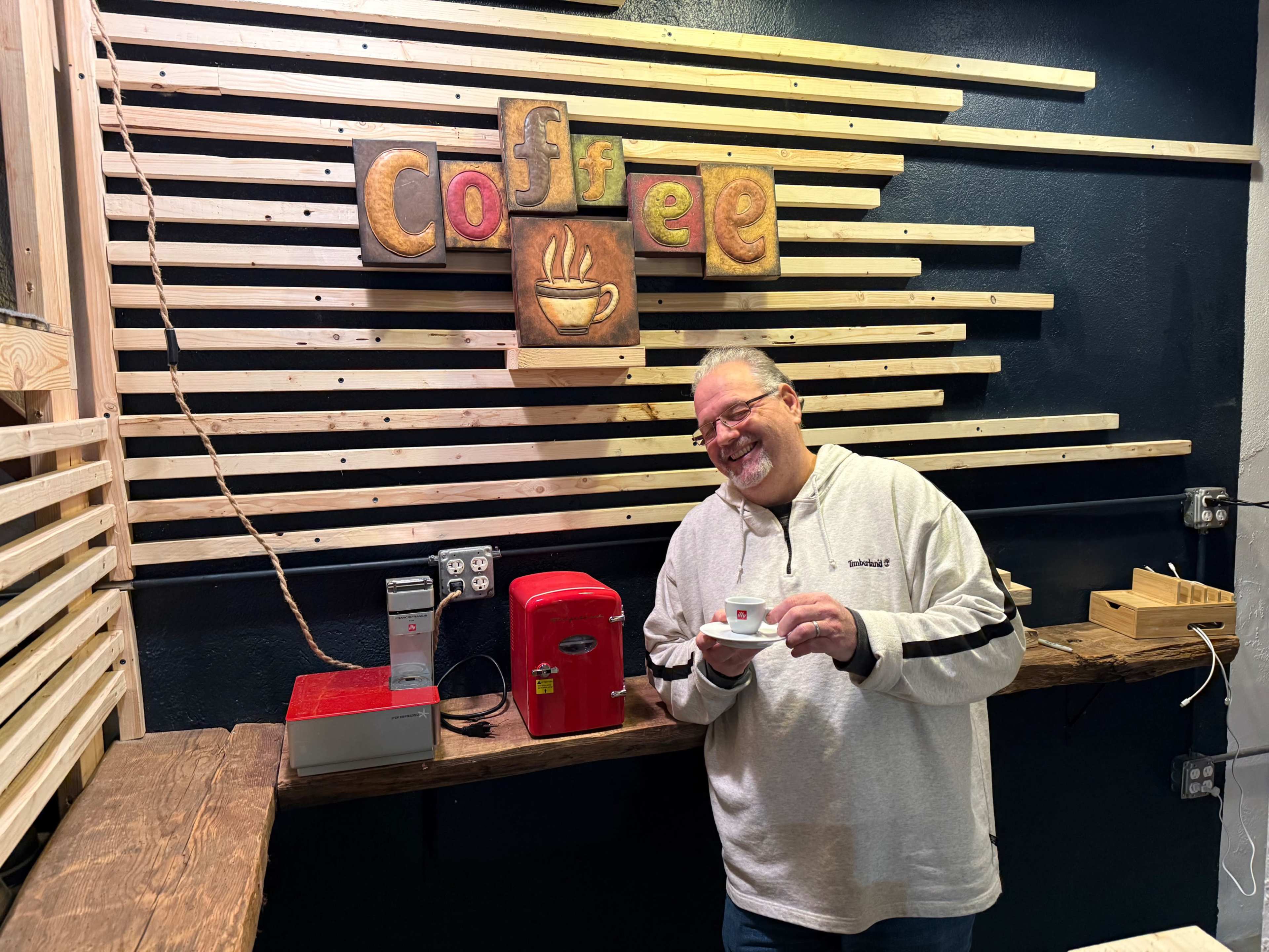 A man holding a small cup stands in front of a wooden coffee-themed backdrop featuring a sign that says "coffee."