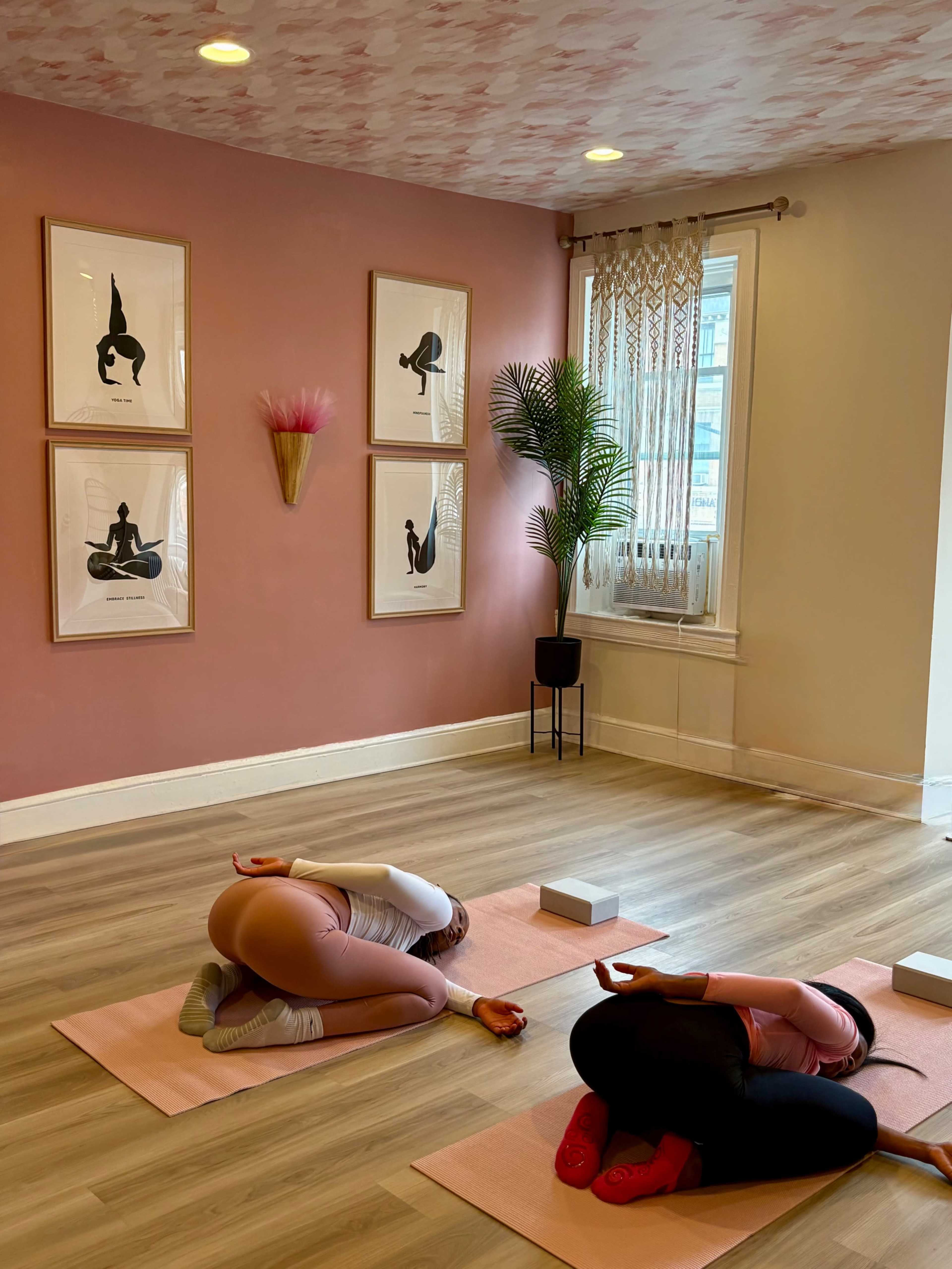 Two individuals are practicing yoga in a warmly lit studio with pink walls and framed artwork, using yoga mats and blocks.