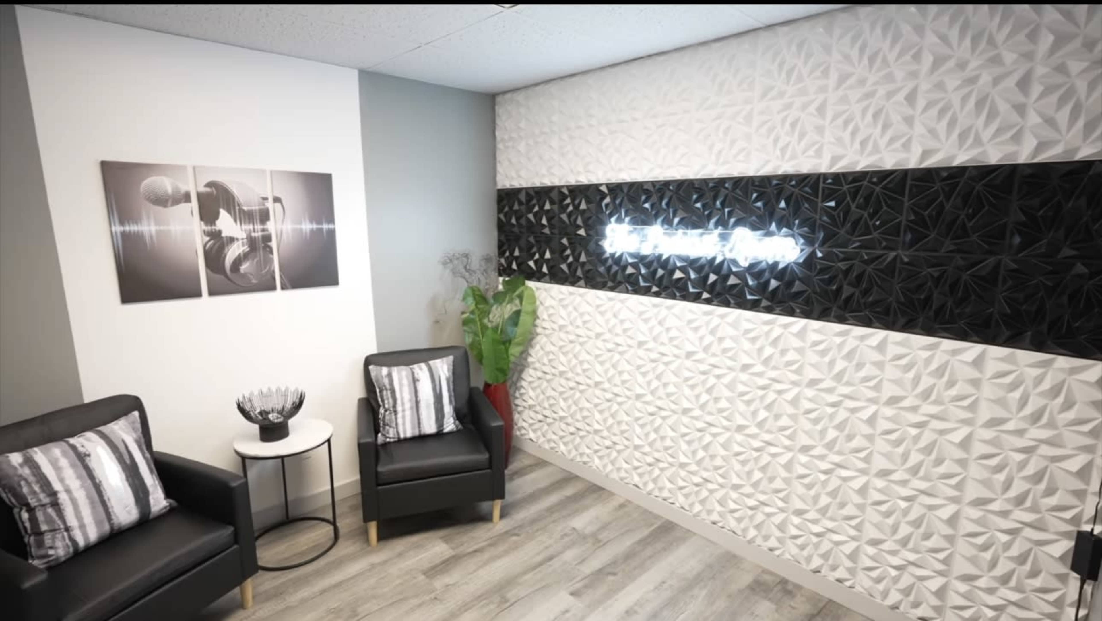 The image shows a modern interior space featuring two black chairs, a small round table with a decorative bowl, and a textured wall with illuminated text.