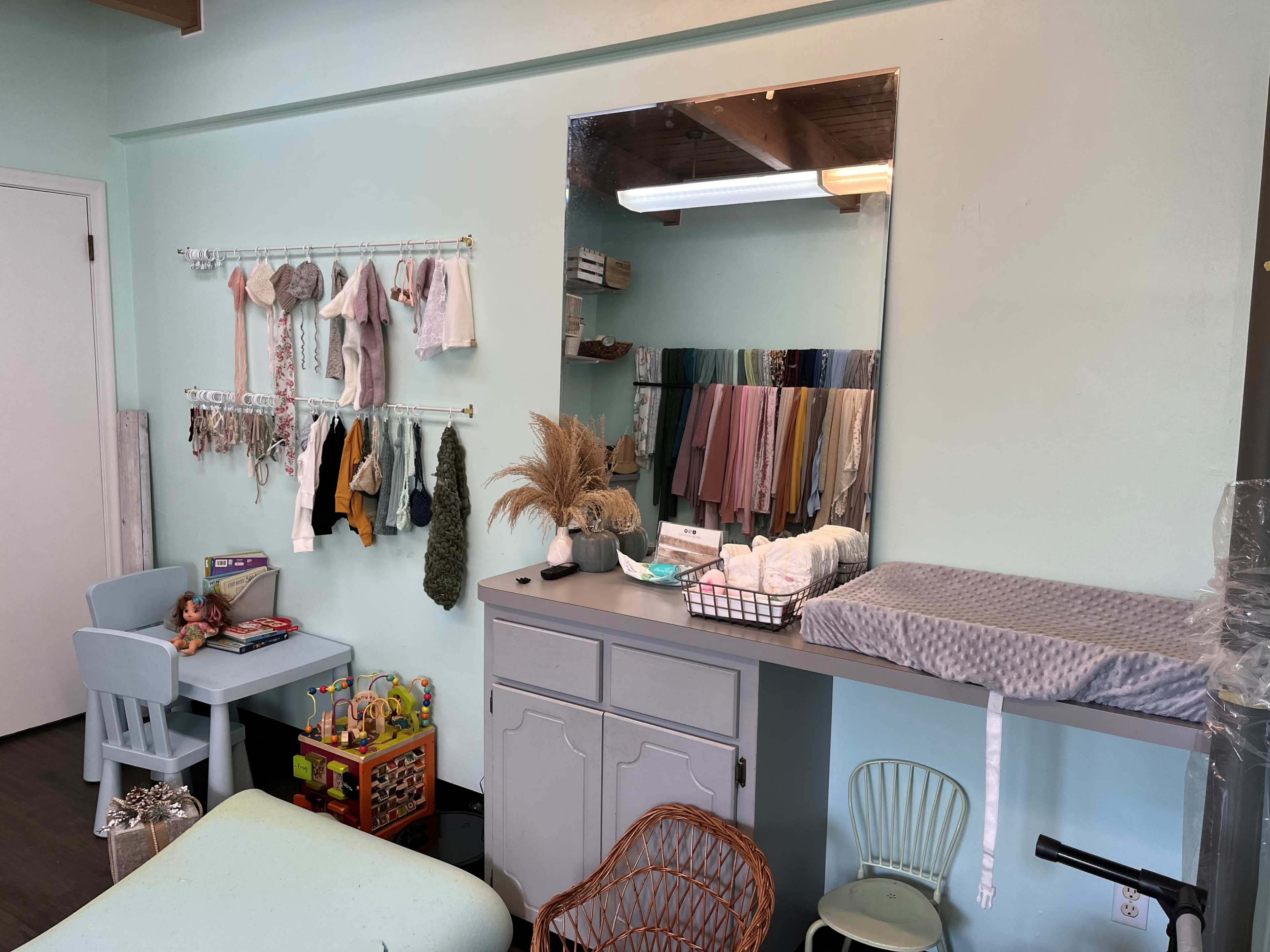 The image shows a light blue room with a changing table, a mirror, various clothes hanging on a rack, and children's toys in a corner.