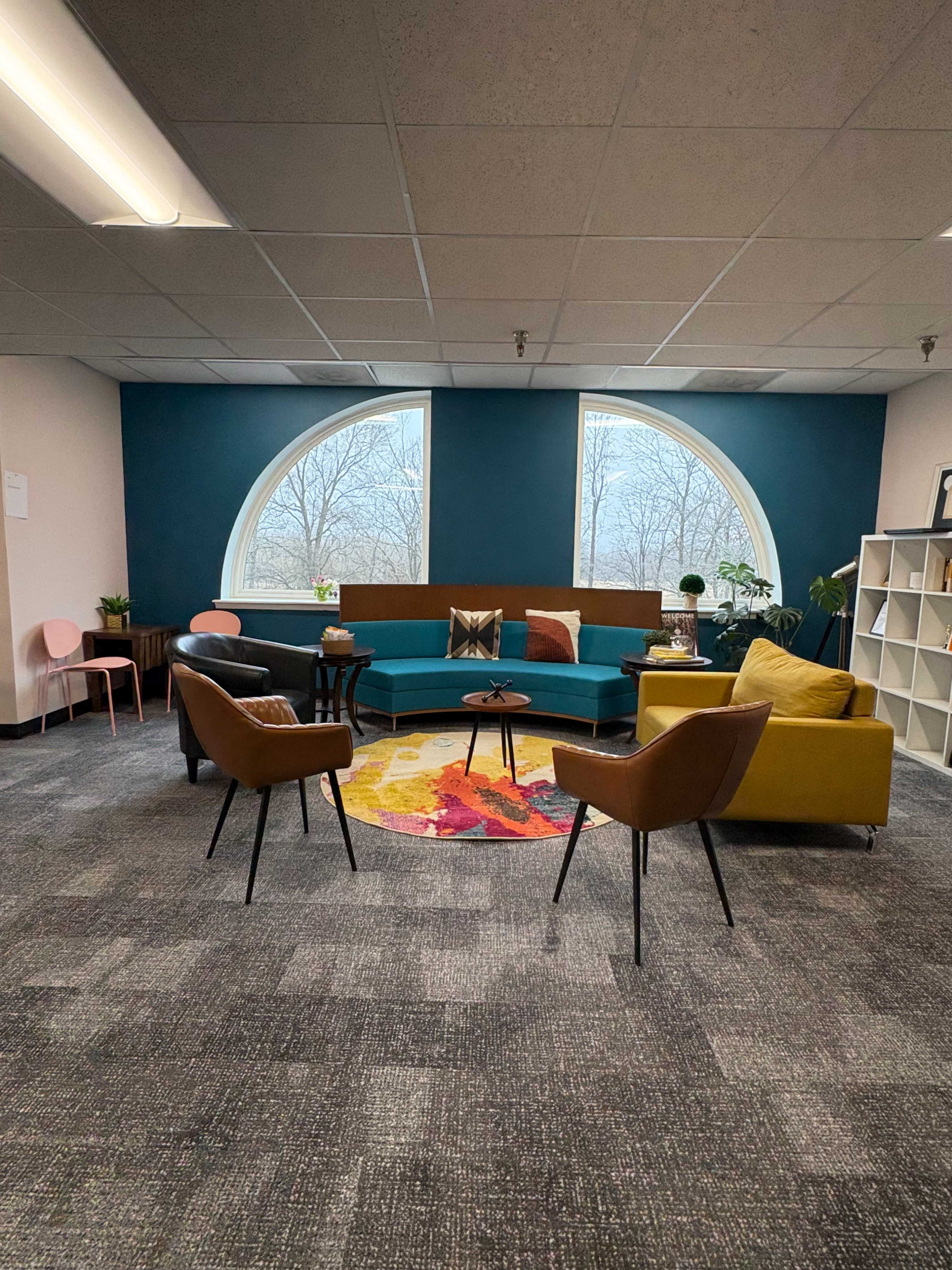 The image shows a modern office lounge area featuring a curved sofa with colorful cushions, a round rug, and various plants, along with a bookshelf and chairs arranged around a central coffee table.