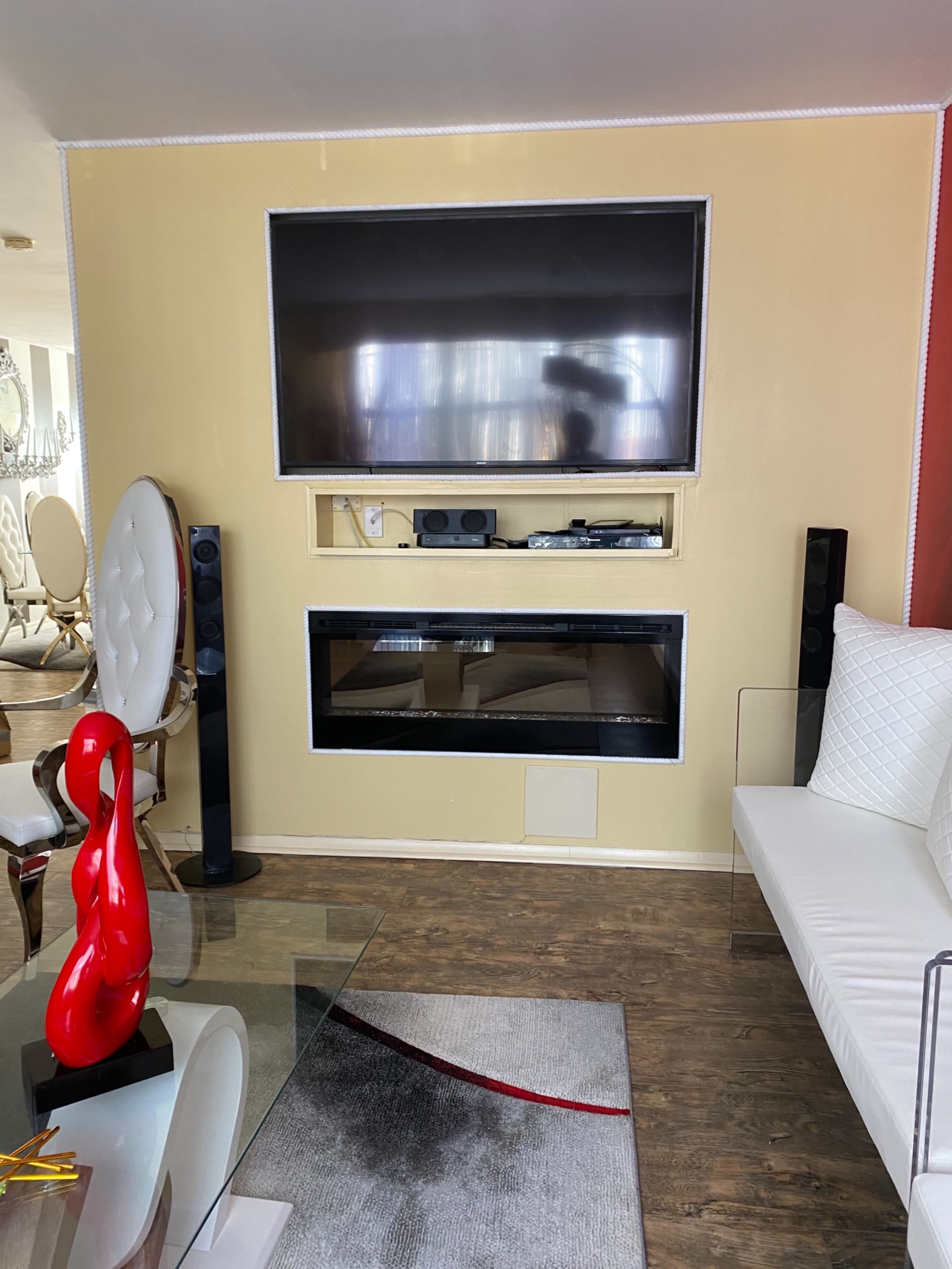 A living room with a mounted television above a modern electric fireplace, flanked by black speakers, a glass coffee table, and white furniture.