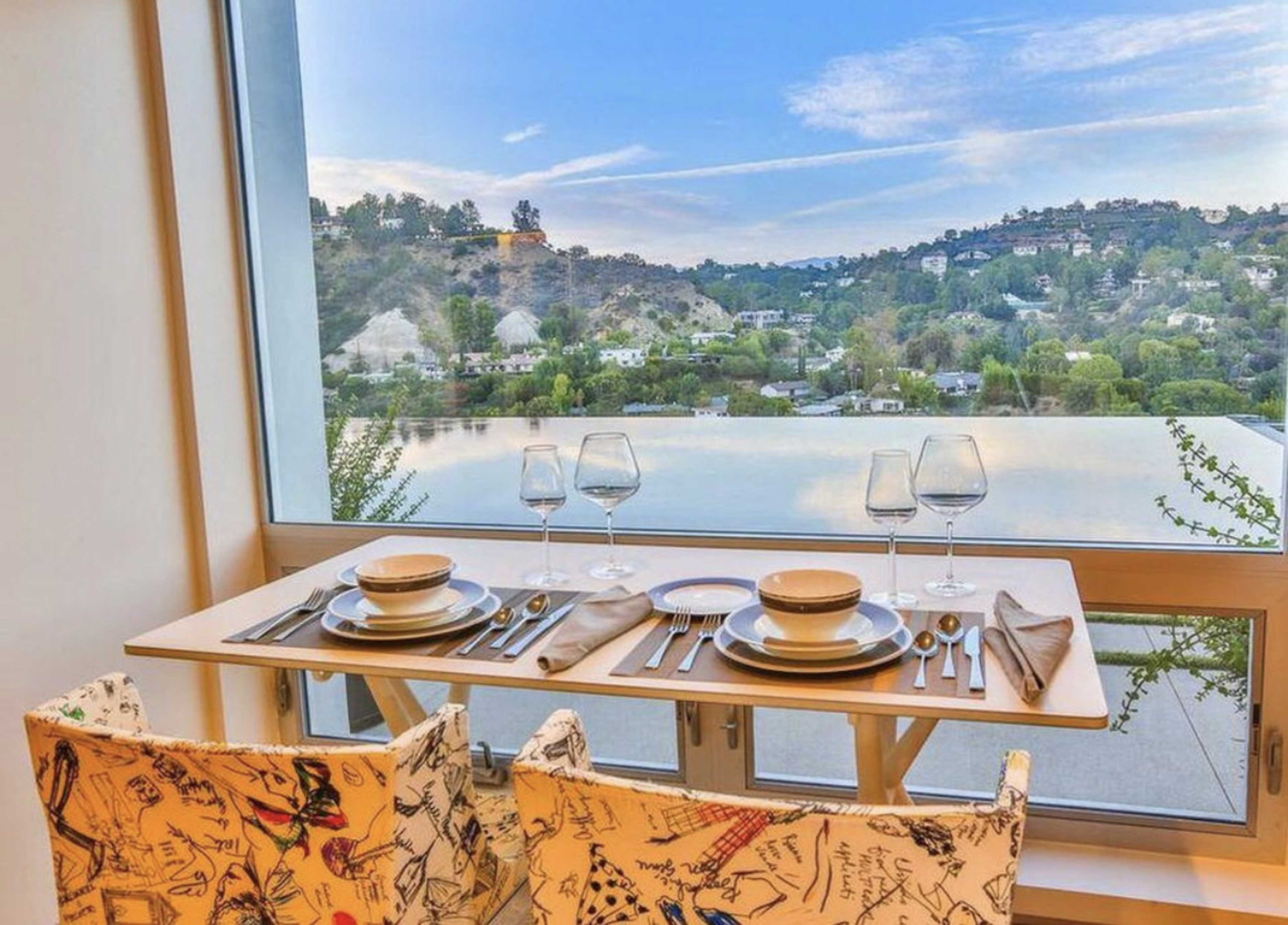 A dining table set for two is positioned by a large window, overlooking a scenic view of hills and a swimming pool.