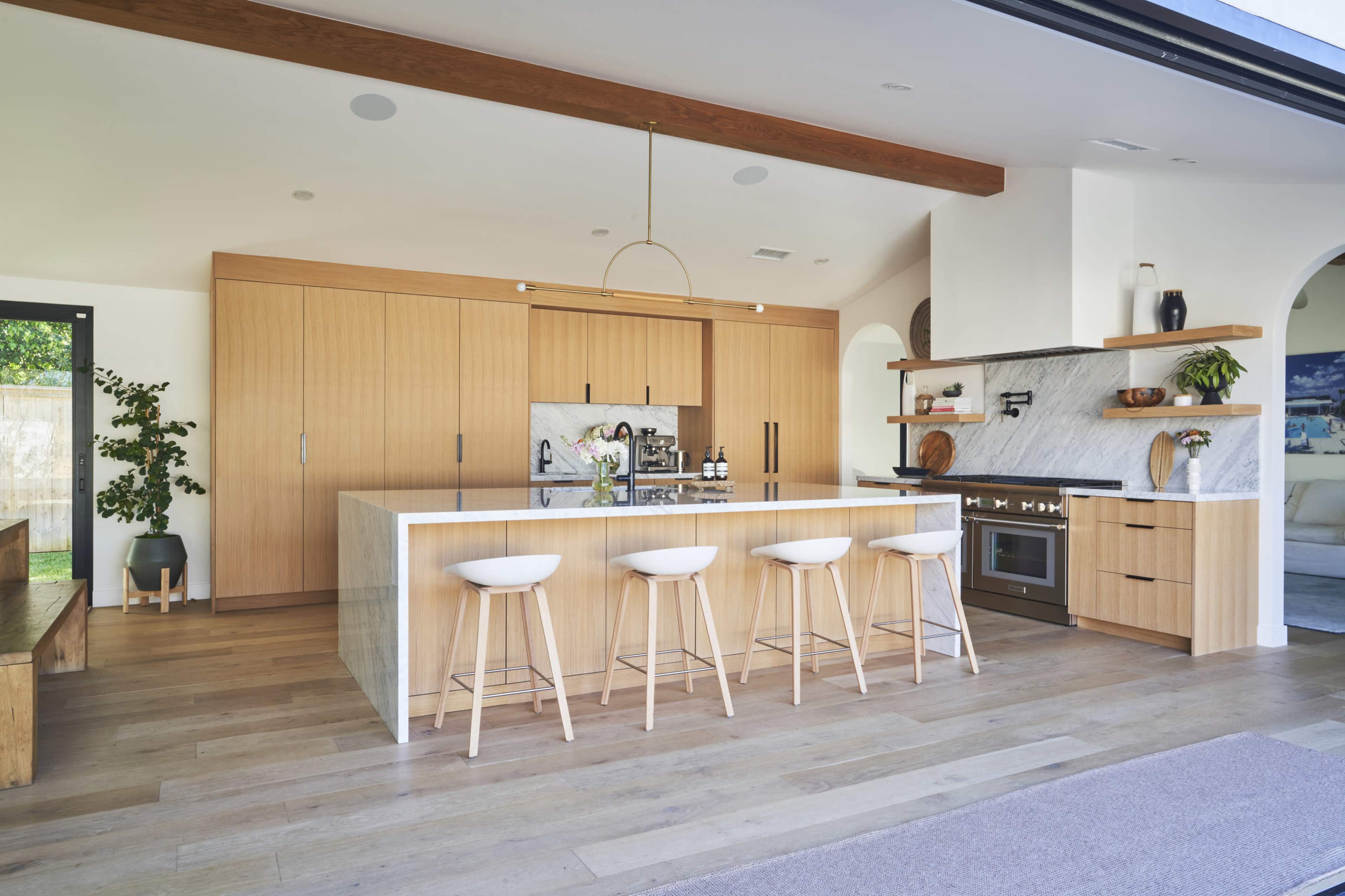 A modern kitchen featuring a large island with four bar stools, wooden cabinetry, marble countertops, and stainless steel appliances.