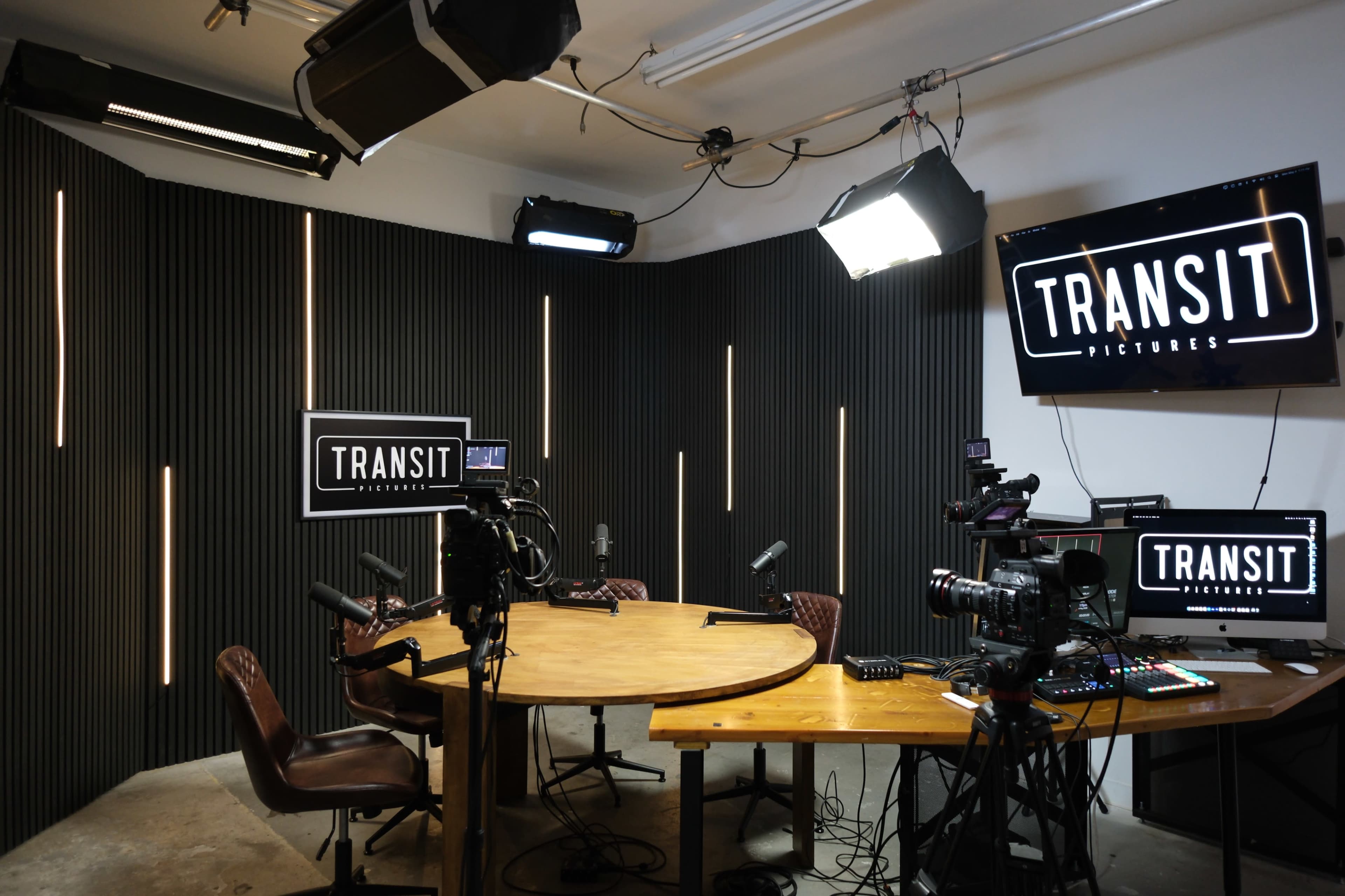 The image shows a modern studio setup for video production, featuring a circular table, several microphones, and multiple cameras, with lighting fixtures and screens displaying the "TRANSIT PICTURES" logo.