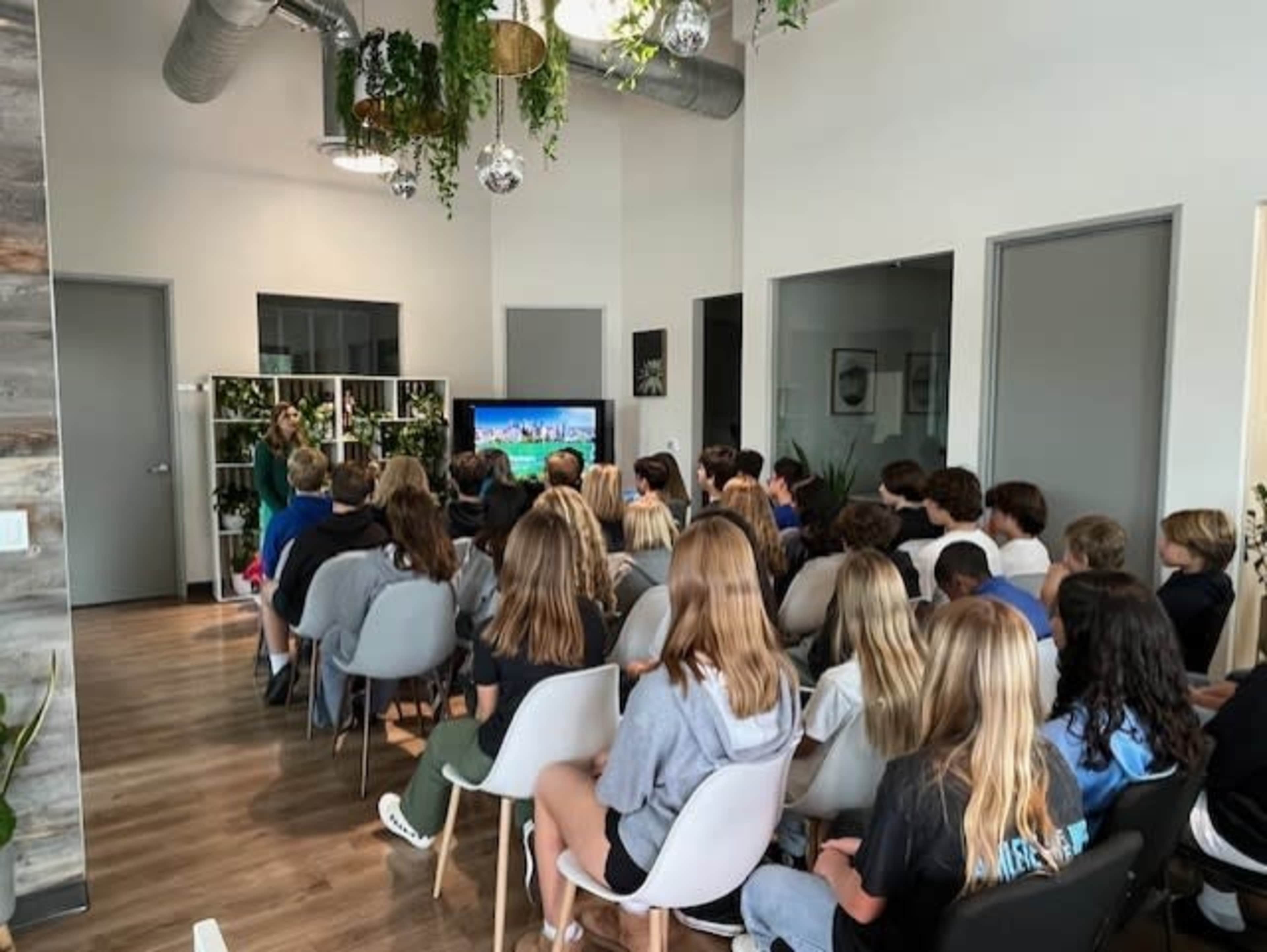 A group of students is seated in a modern classroom, facing a presenter standing near a television screen.