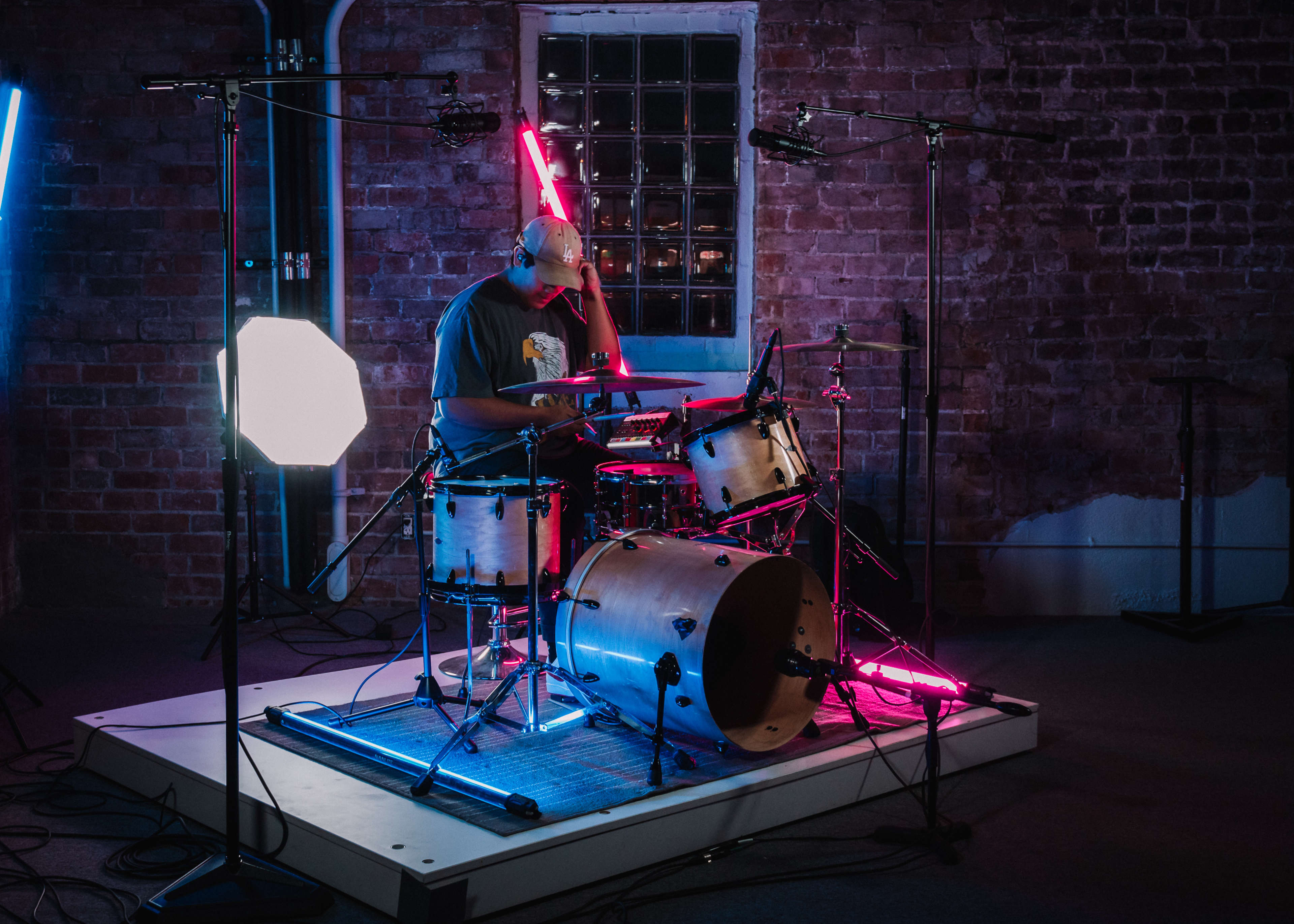 A drummer plays on a well-lit stage surrounded by various microphones and colorful lighting against a brick wall.