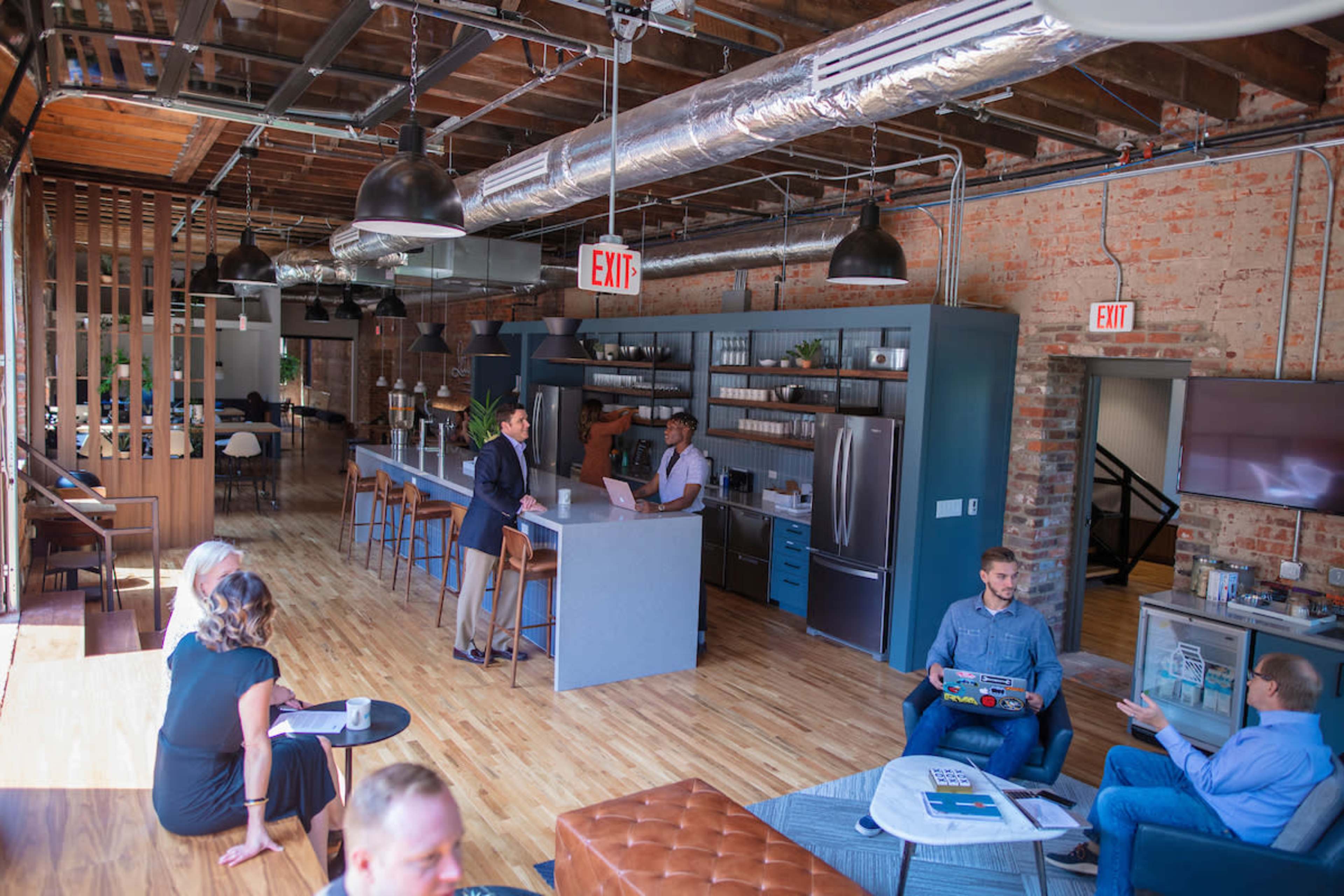 A modern office space features a kitchen area, communal seating, and people engaged in conversation and work activities.