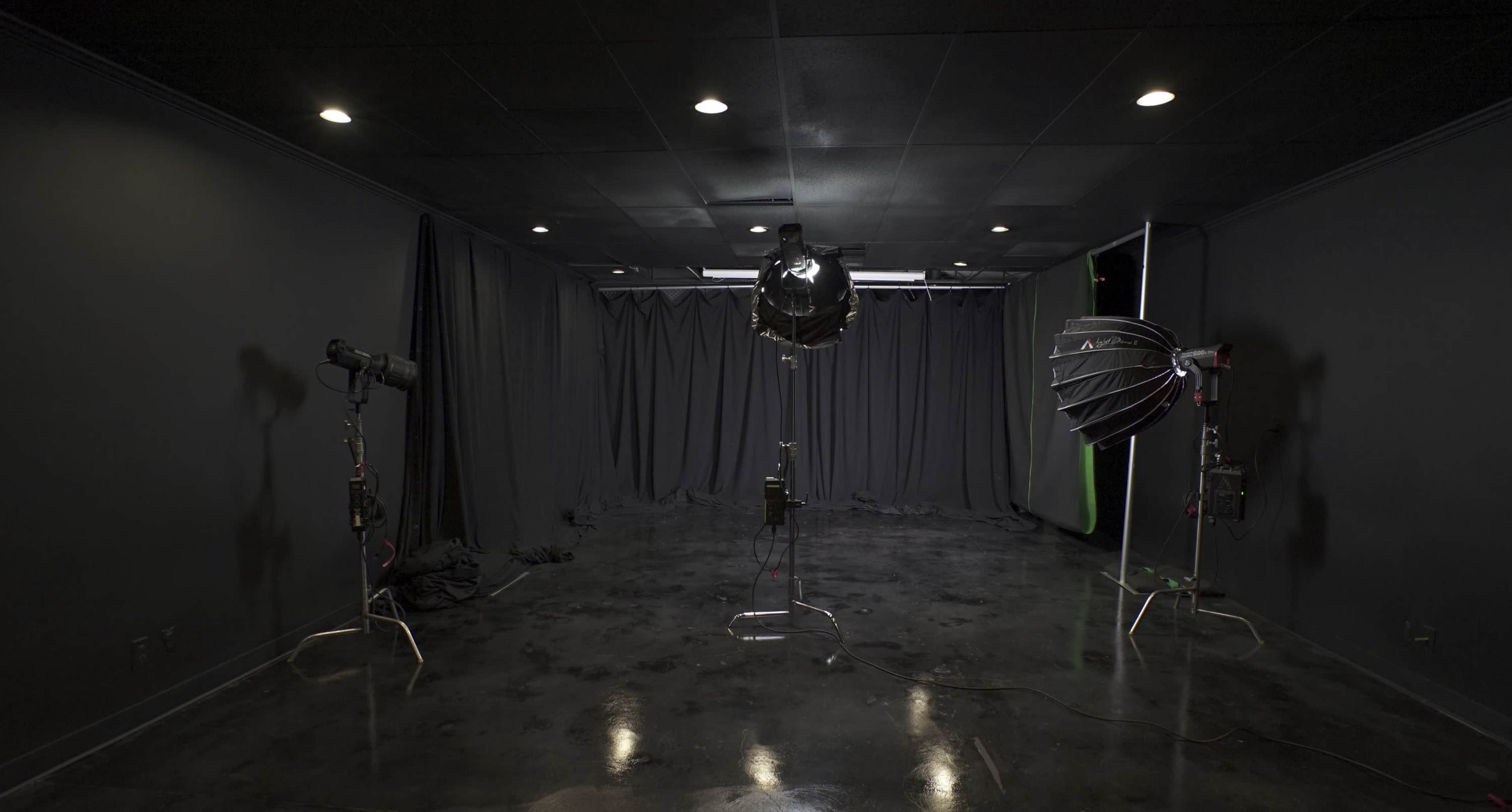 A photography studio with dark walls and floor, featuring two light setups and a backdrop of black curtains.