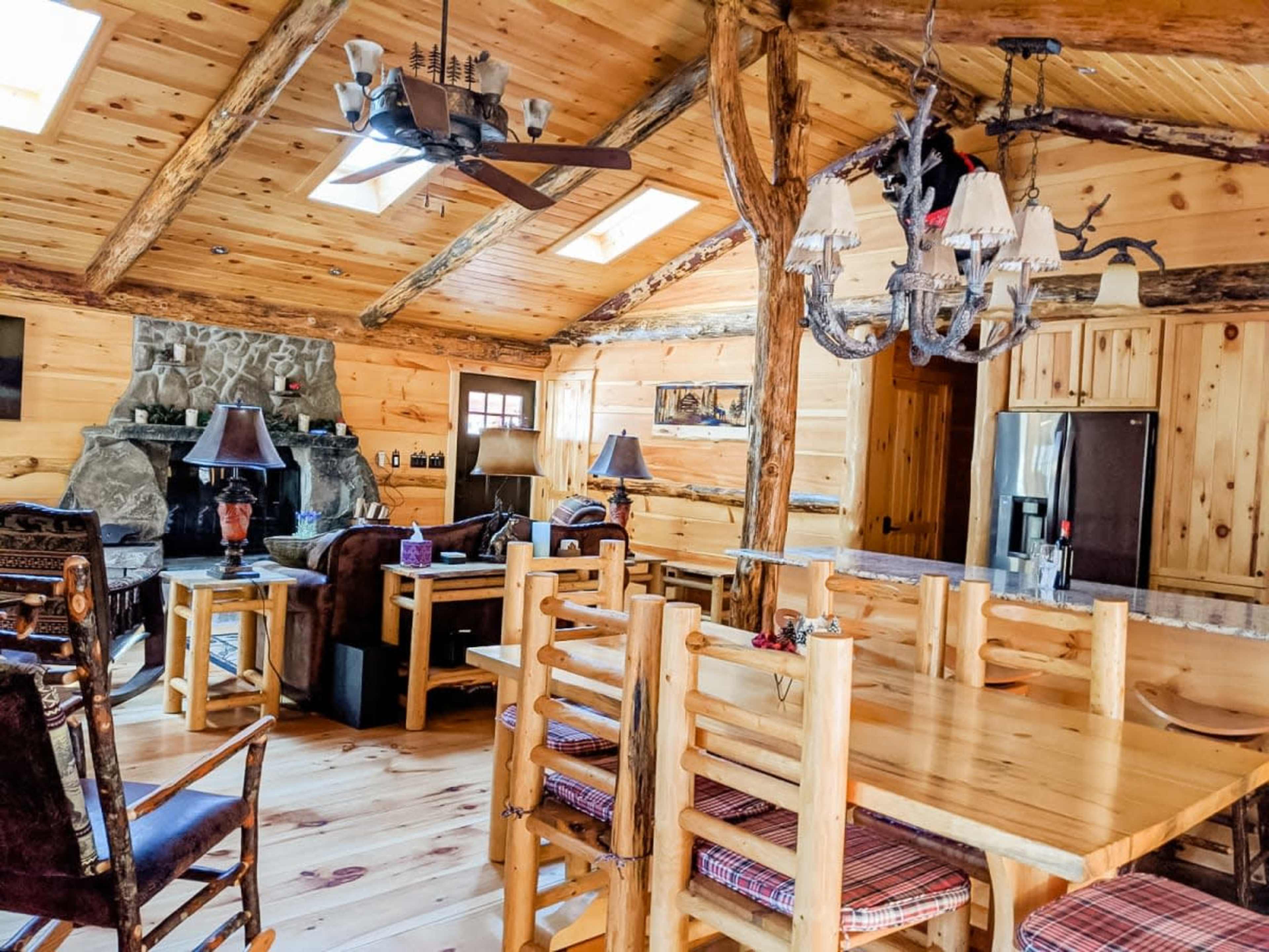 A rustic wooden interior of a cabin with a stone fireplace, dining area, and open-beamed ceilings.