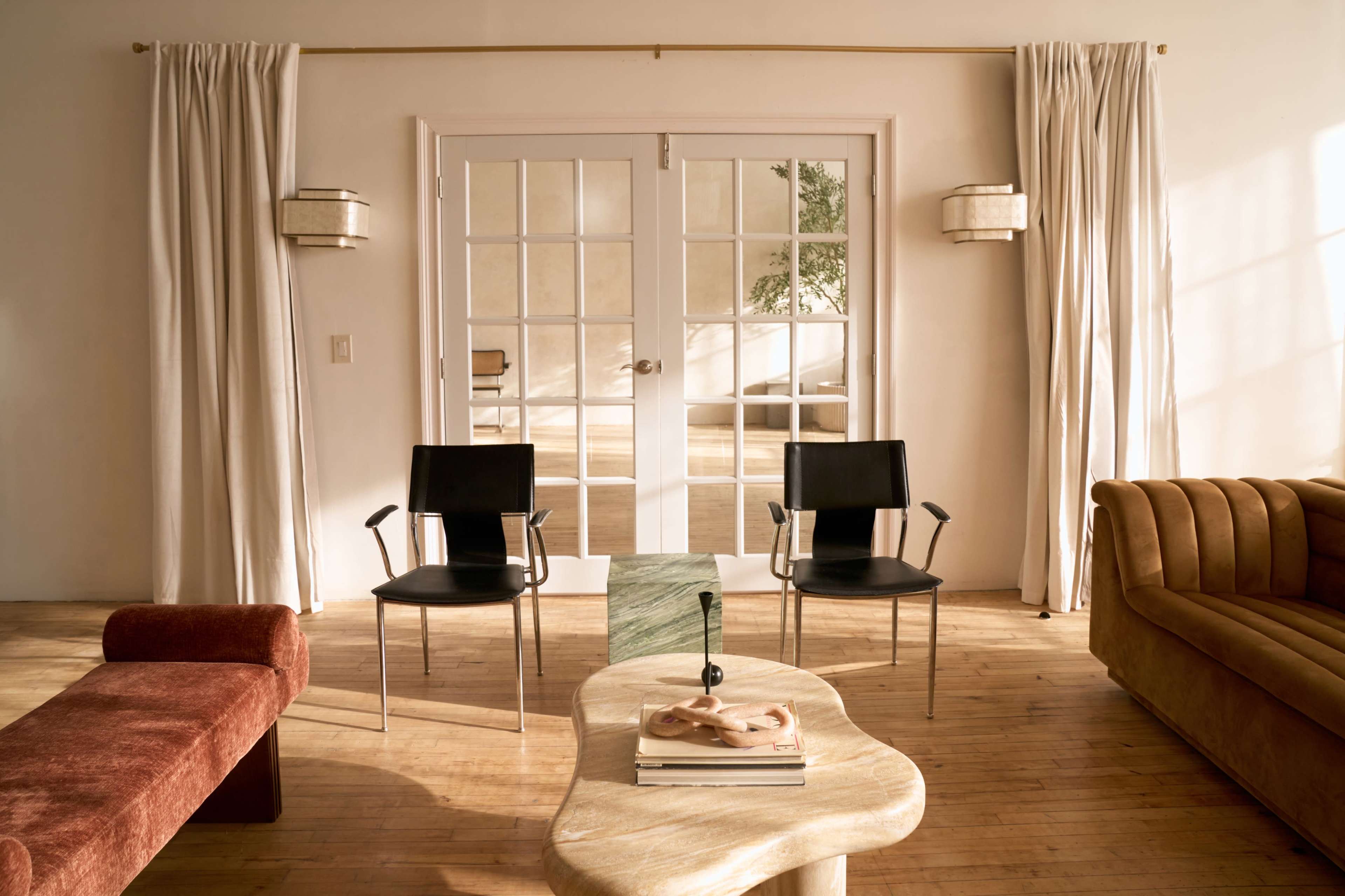The image shows a spacious, well-lit room featuring two black chairs and a curved wooden coffee table, with large glass doors leading to another area and neutral curtains framing the windows.