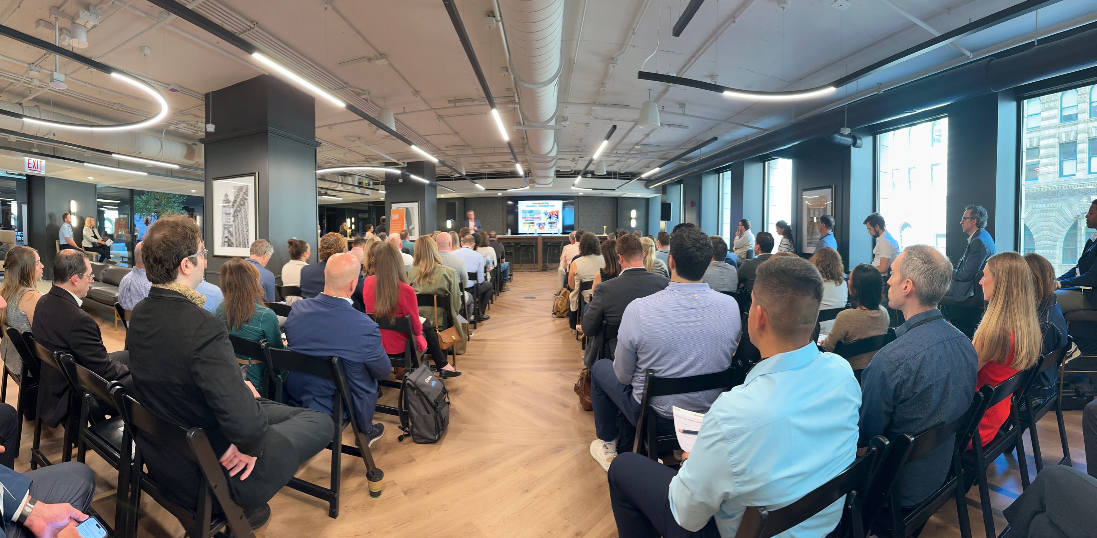 A large audience is seated in a modern conference room, attentively facing a presentation screen at the front of the space.