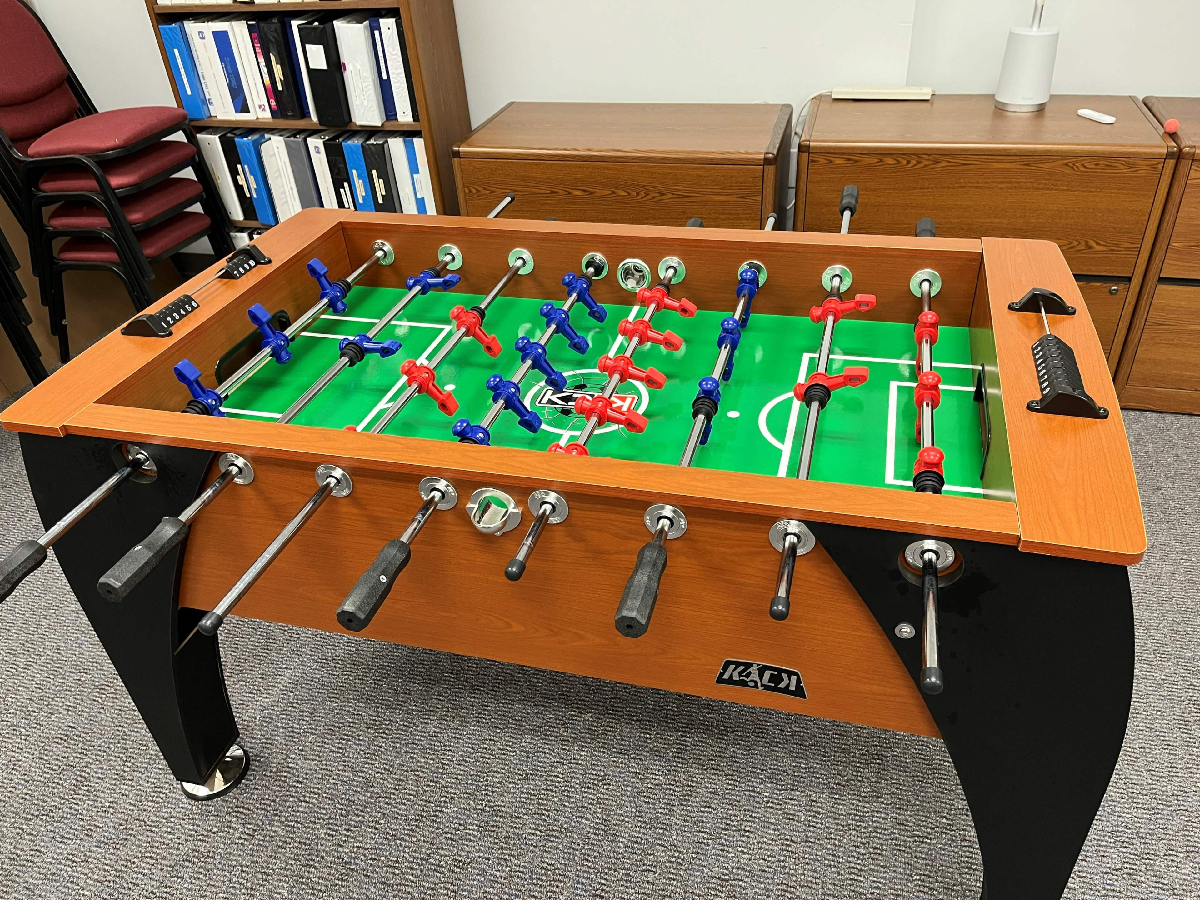 A foosball table with red and blue players is positioned in an office setting, surrounded by chairs and storage cabinets.