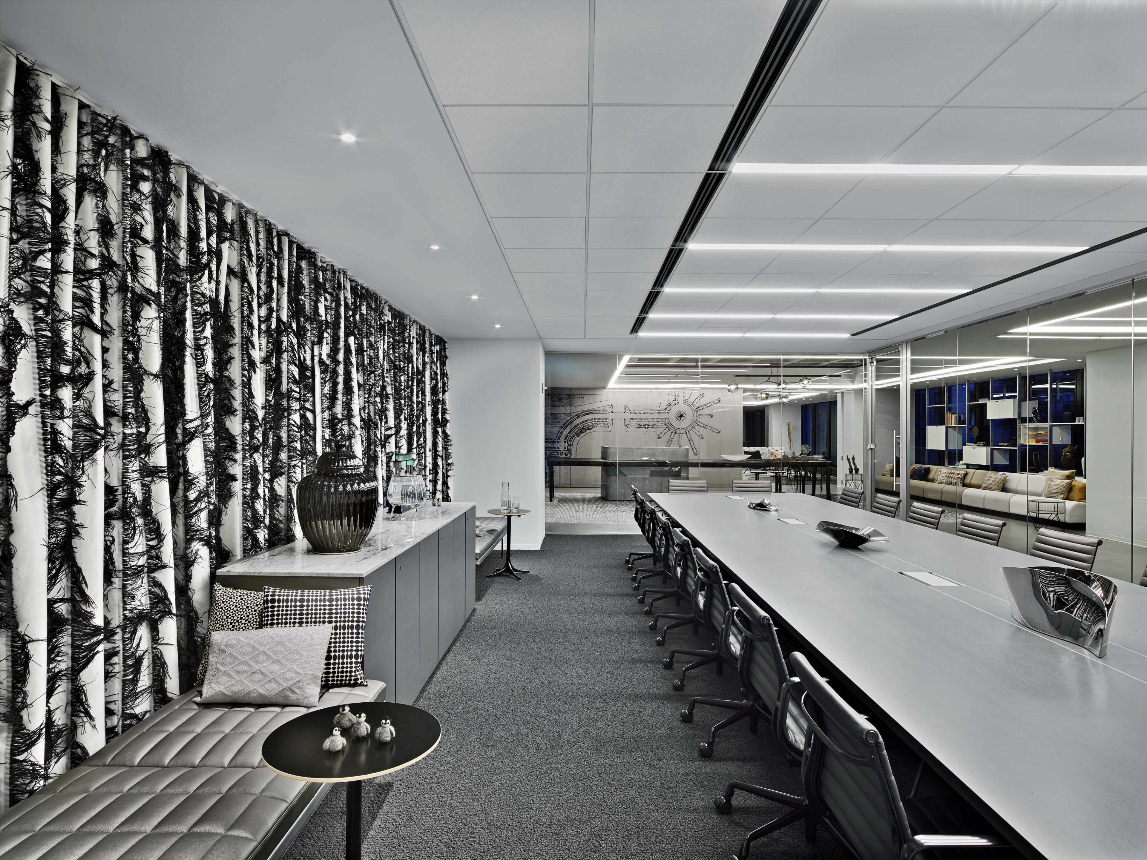 A modern conference room features a long table with black chairs, a textured wall adorned with a tree design, and a glass-walled area displaying additional seating.