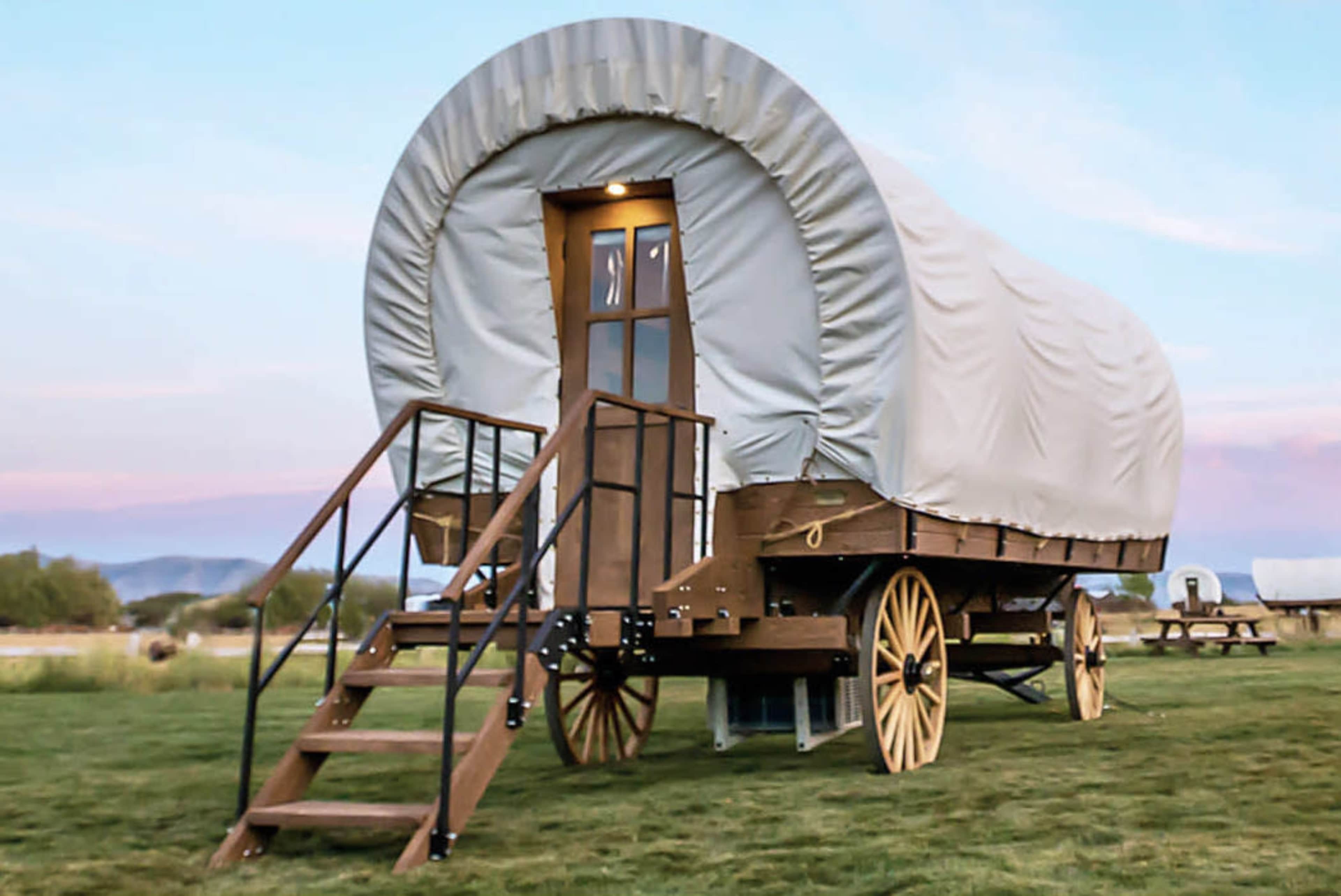 A covered wagon with wooden wheels and a staircase is set on a grassy landscape under a pastel sky.