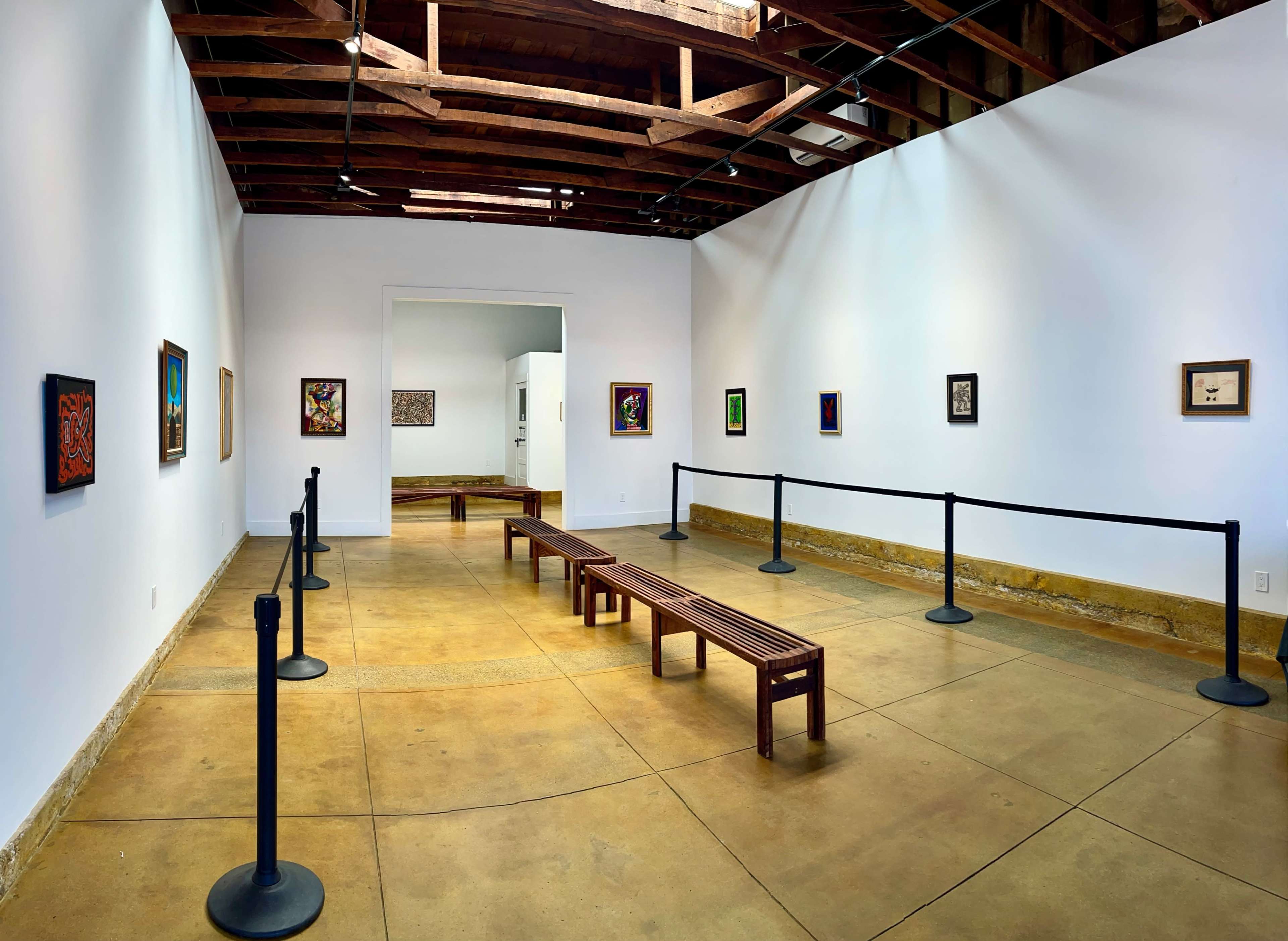 A spacious art gallery features wooden benches and various framed artworks displayed on plain white walls.