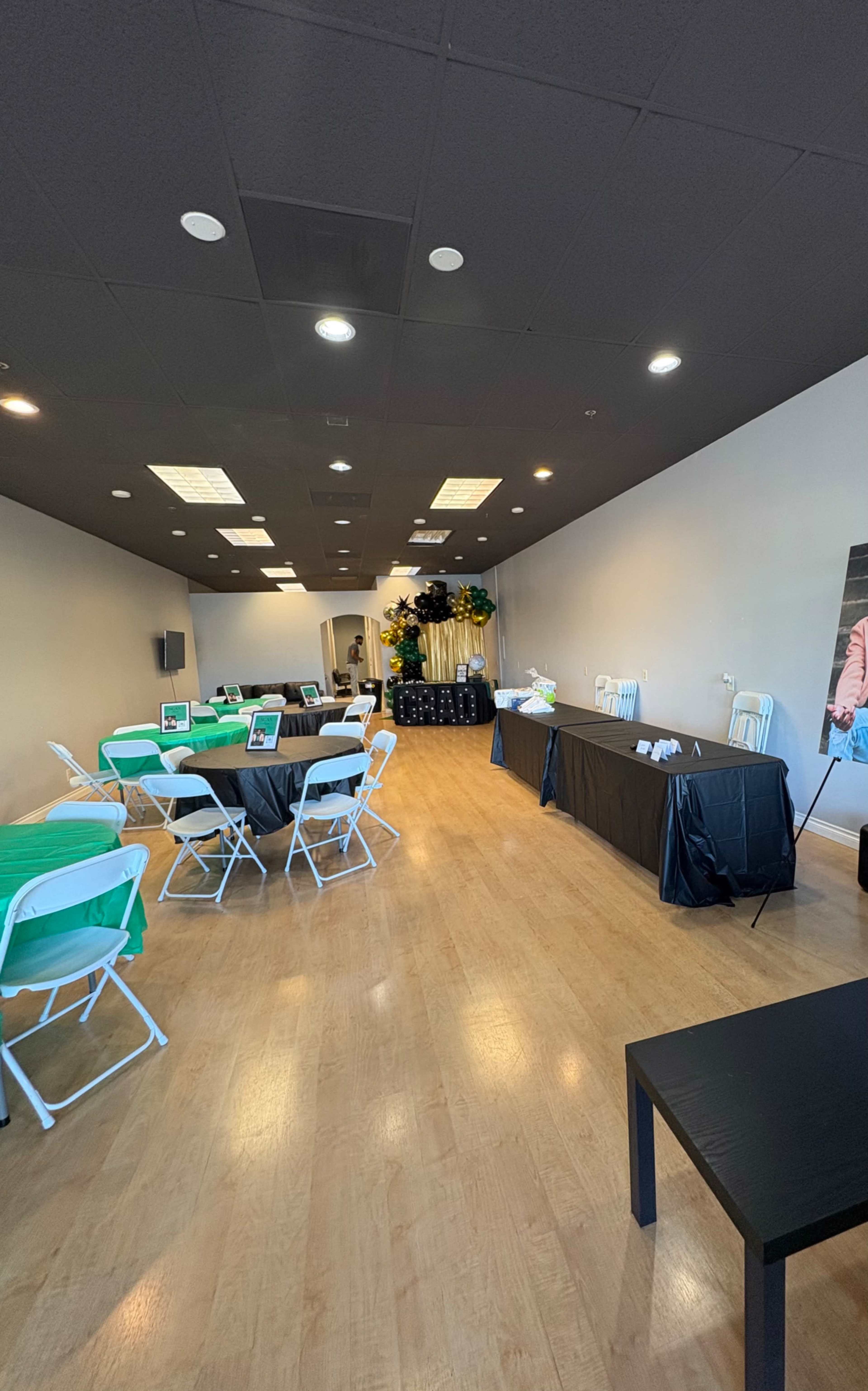 The image shows a spacious event room with tables and chairs set up, a black table at the back, and decorative plants in the corner.
