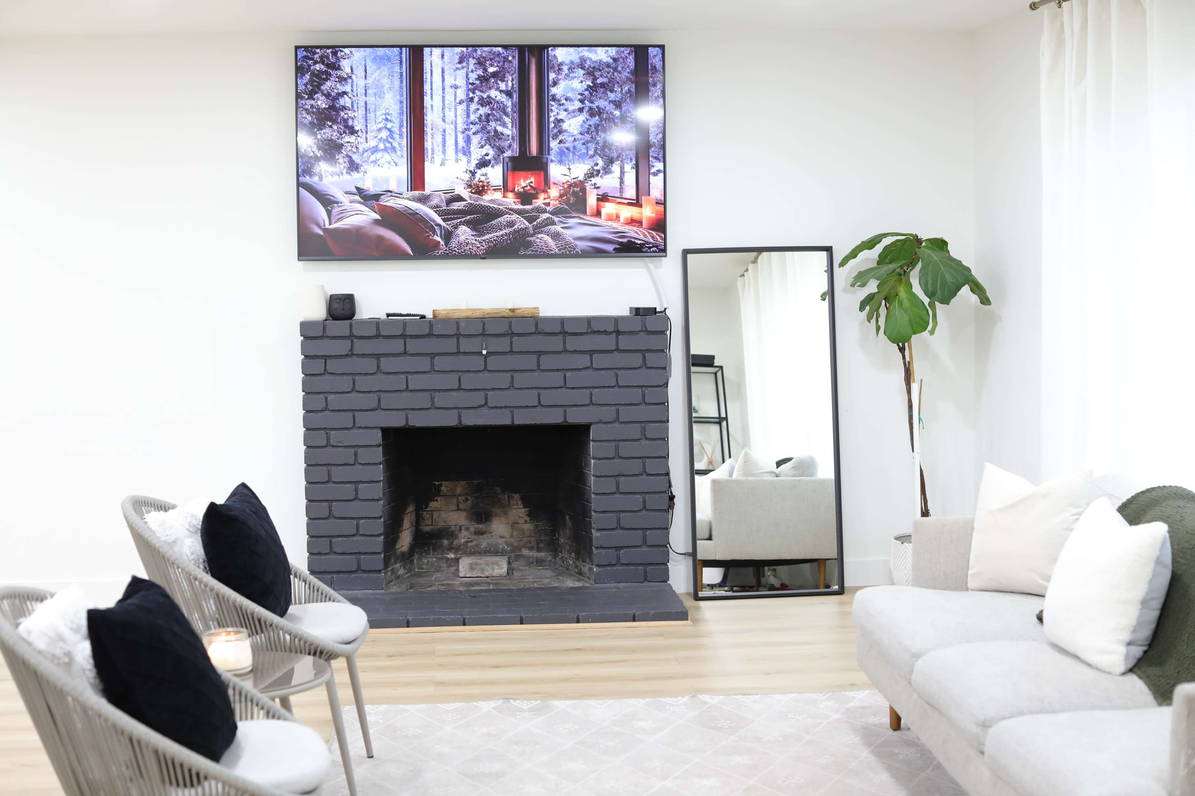 A cozy living room features a dark brick fireplace, a large television mounted above it, a mirror beside the seating area, and two stylish chairs with black cushions.
