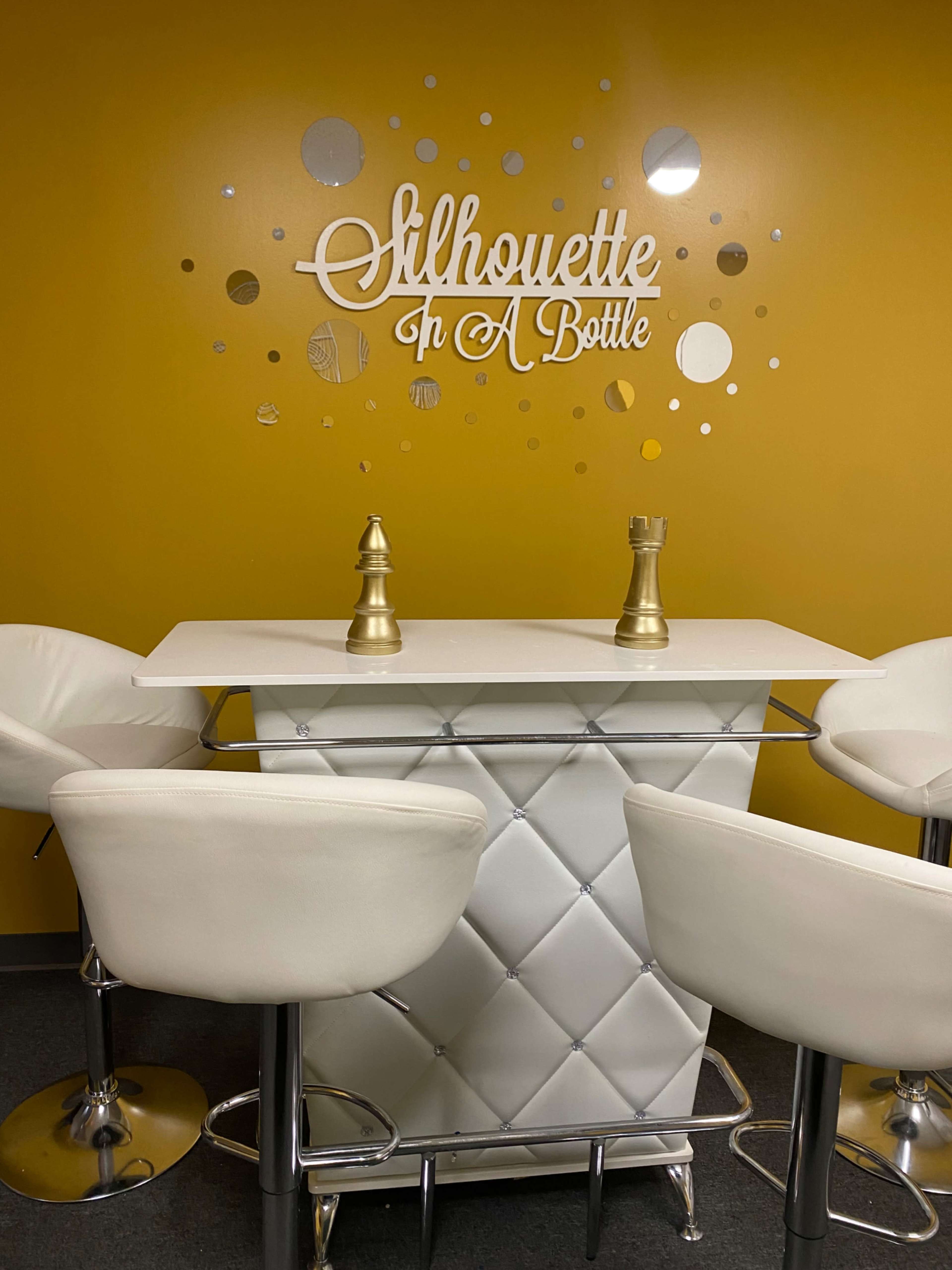 A white bar table with decorative gold accents is surrounded by four white bar stools against a yellow wall featuring the words "Silhouette In A Bottle."