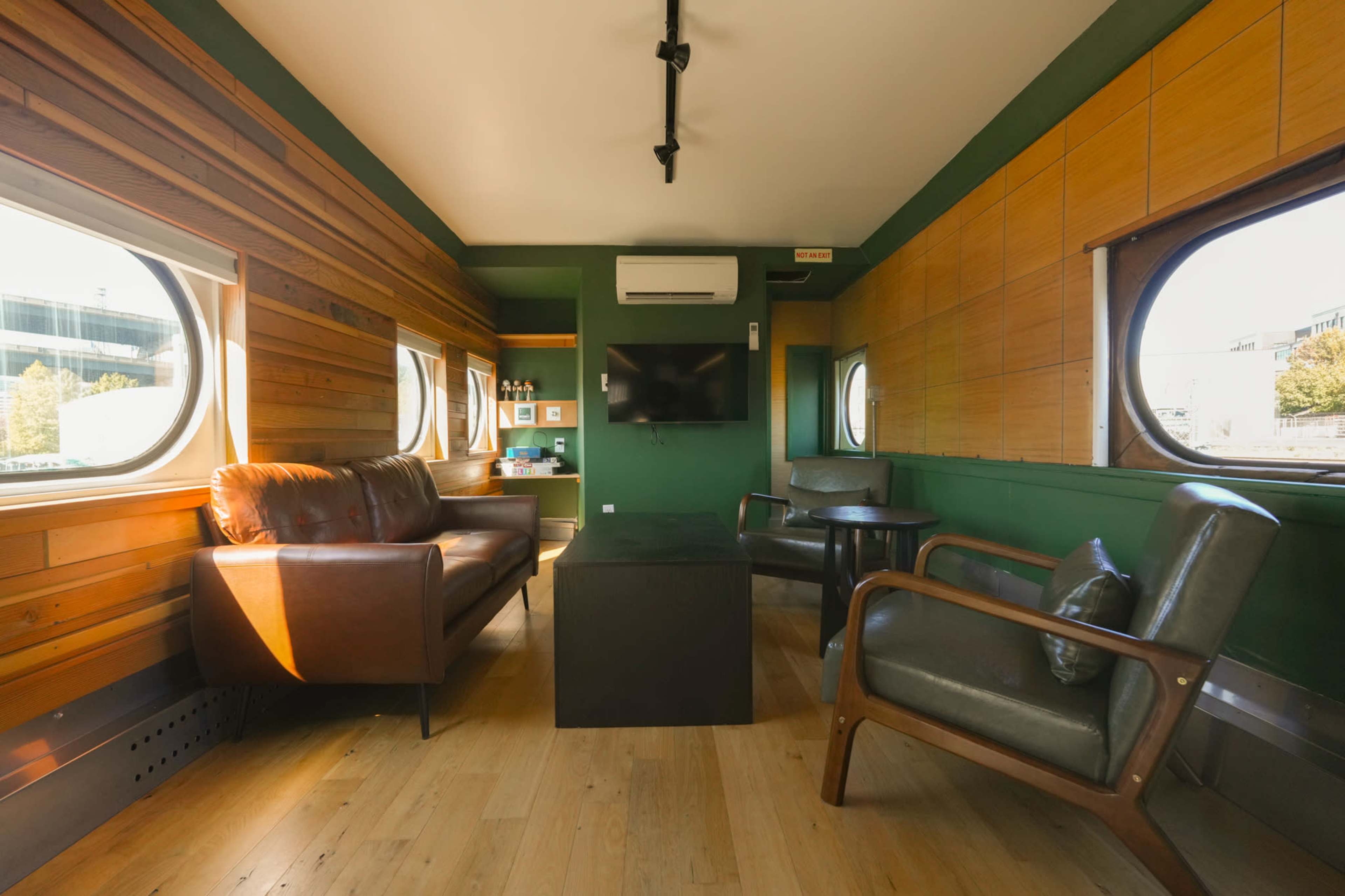The image shows a modern interior of a room with wooden walls, large round windows, a leather sofa, and a small table in the center.