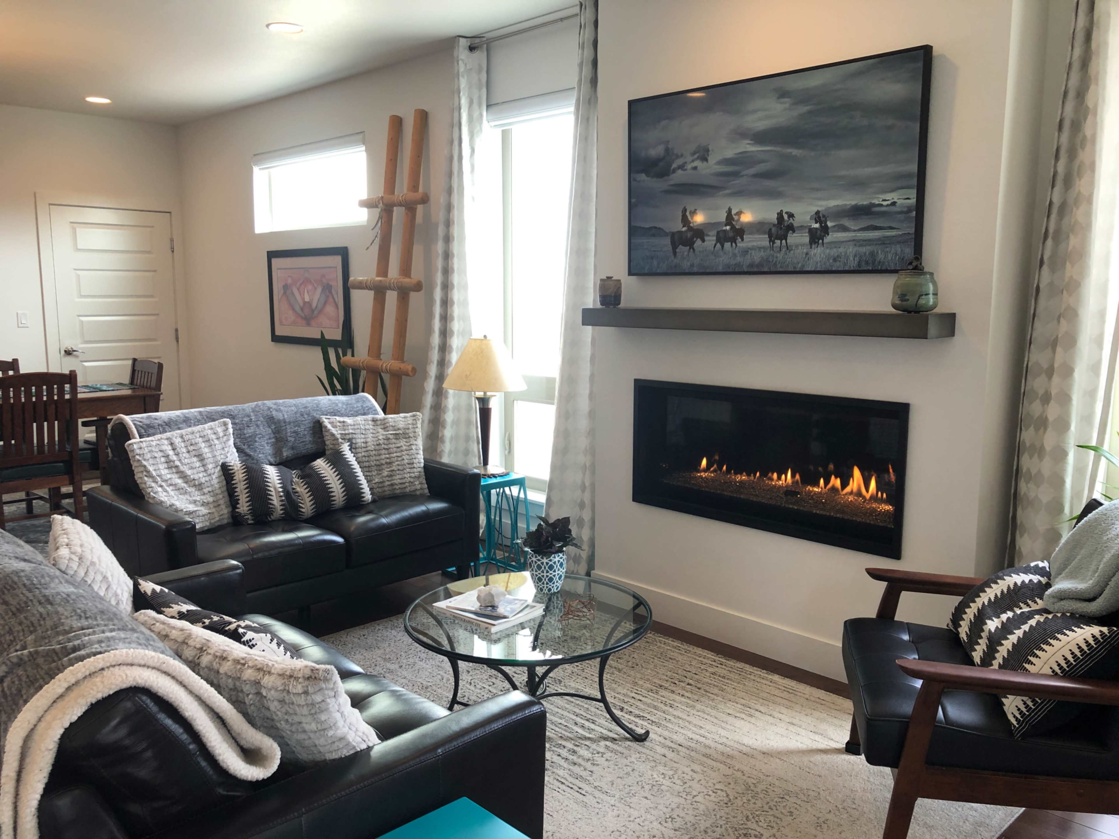 A modern living room features a black leather sofa, a glass coffee table, a fireplace with a mantel, and large windows that let in natural light.