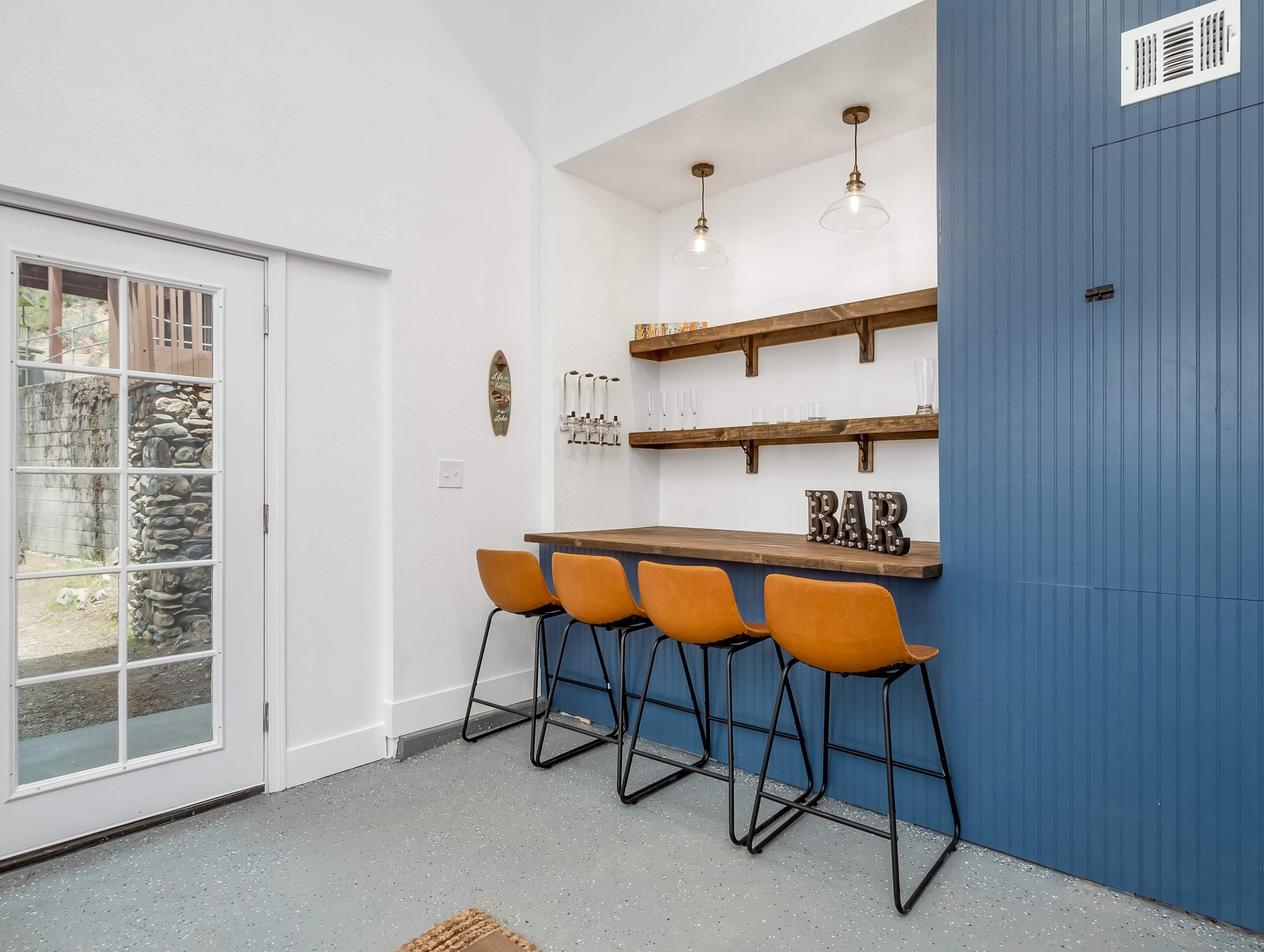 A modern bar area with three orange stools, a wooden countertop, a blue wall with shelves, and a glass door leading outside.