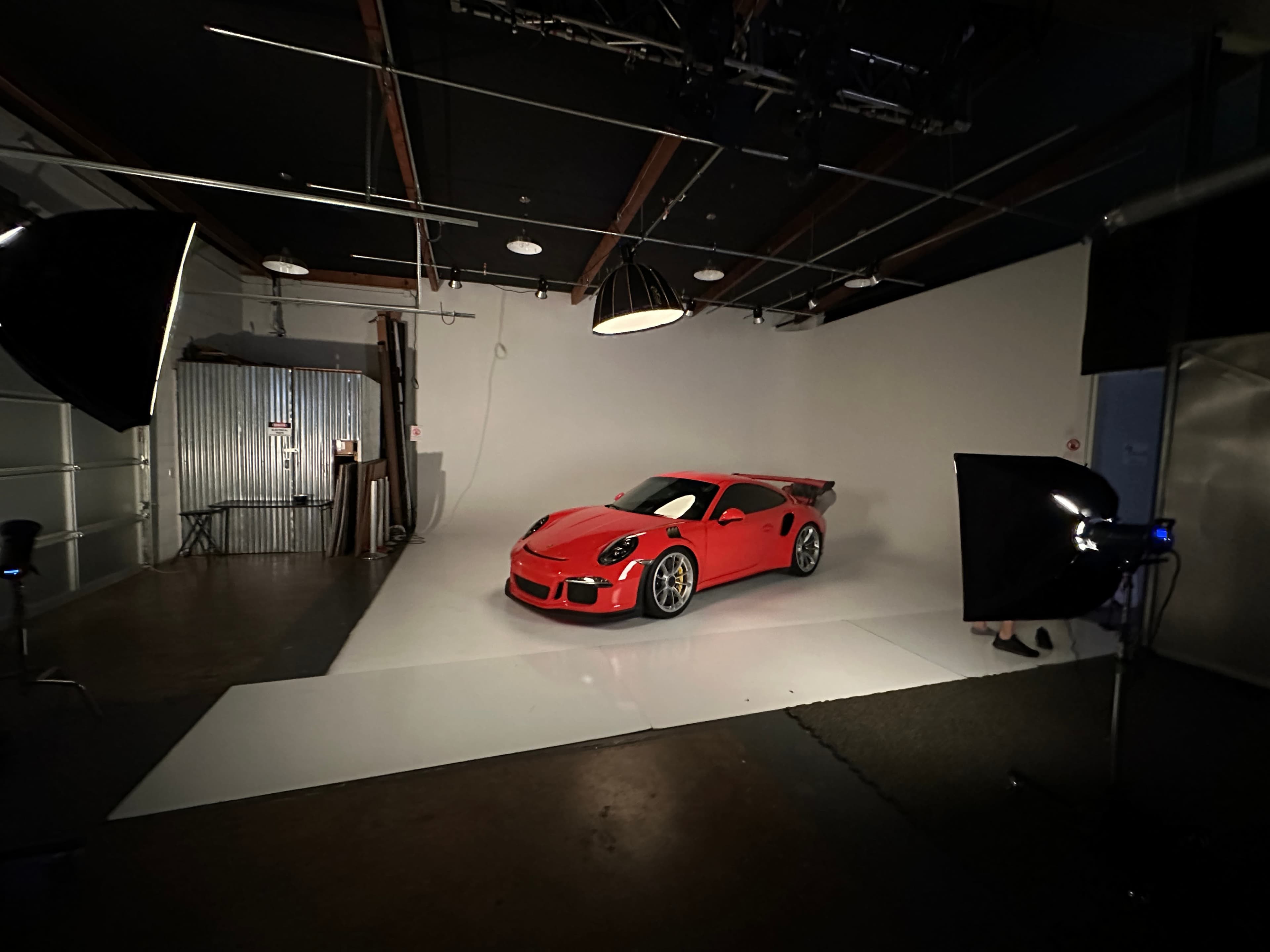 A bright red sports car is displayed on a white backdrop in a photography studio, surrounded by professional lighting equipment.