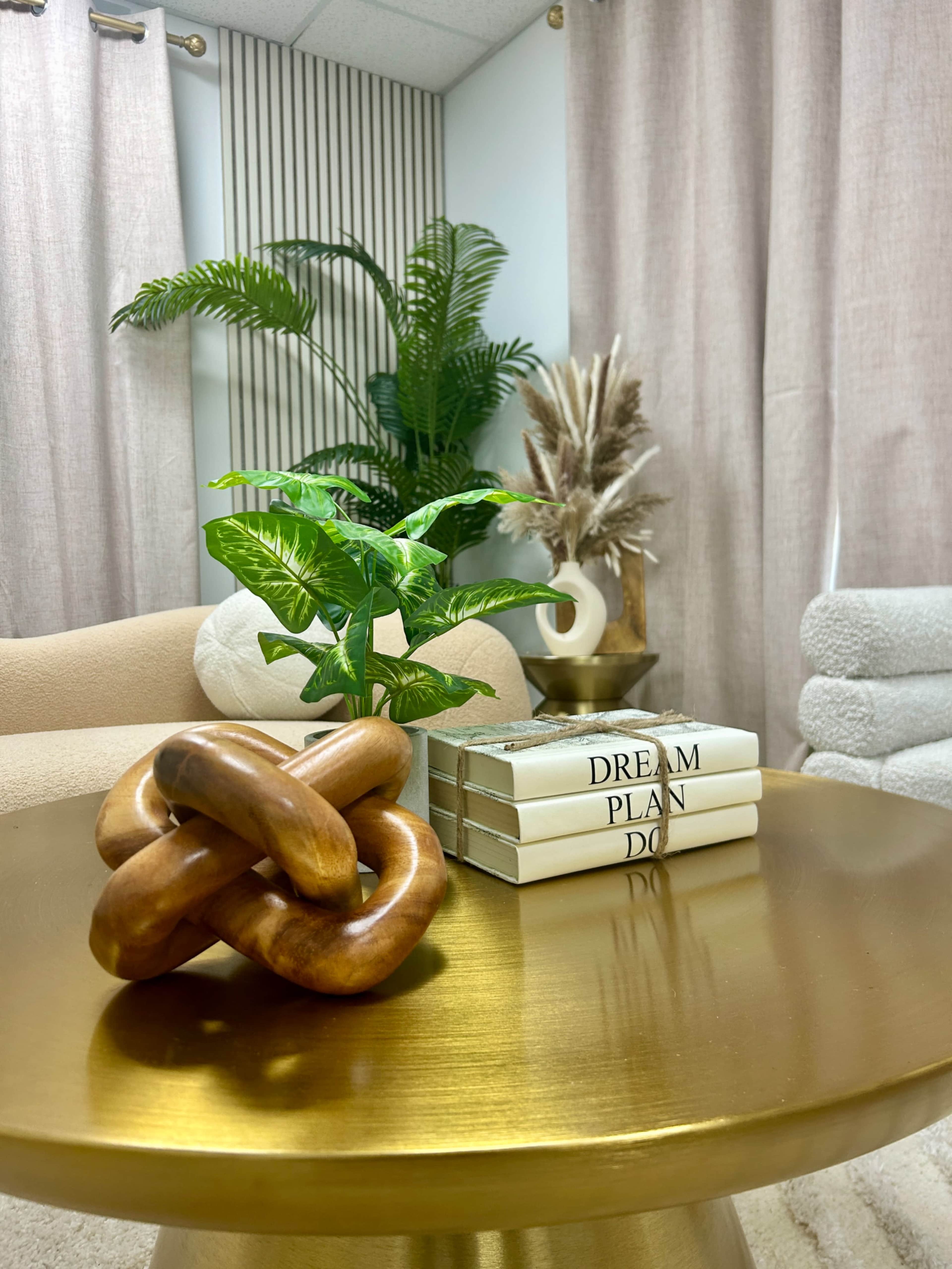 A polished gold table holds a wooden knot sculpture and stacked books, with a small potted plant and decorative elements in the background.