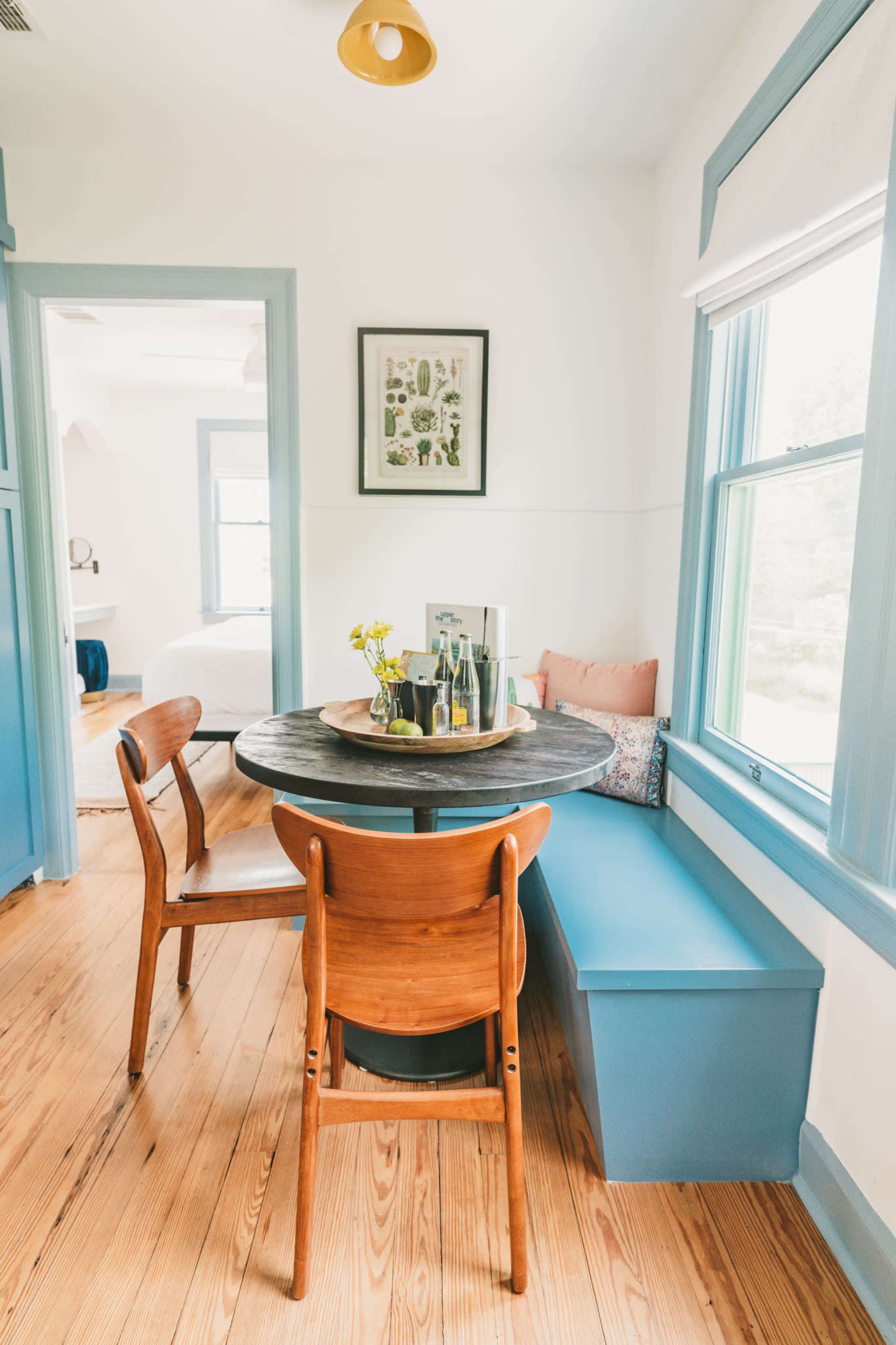 A round table with three wooden chairs and a blue bench is positioned near a window in a bright room with light-colored walls and blue trim.