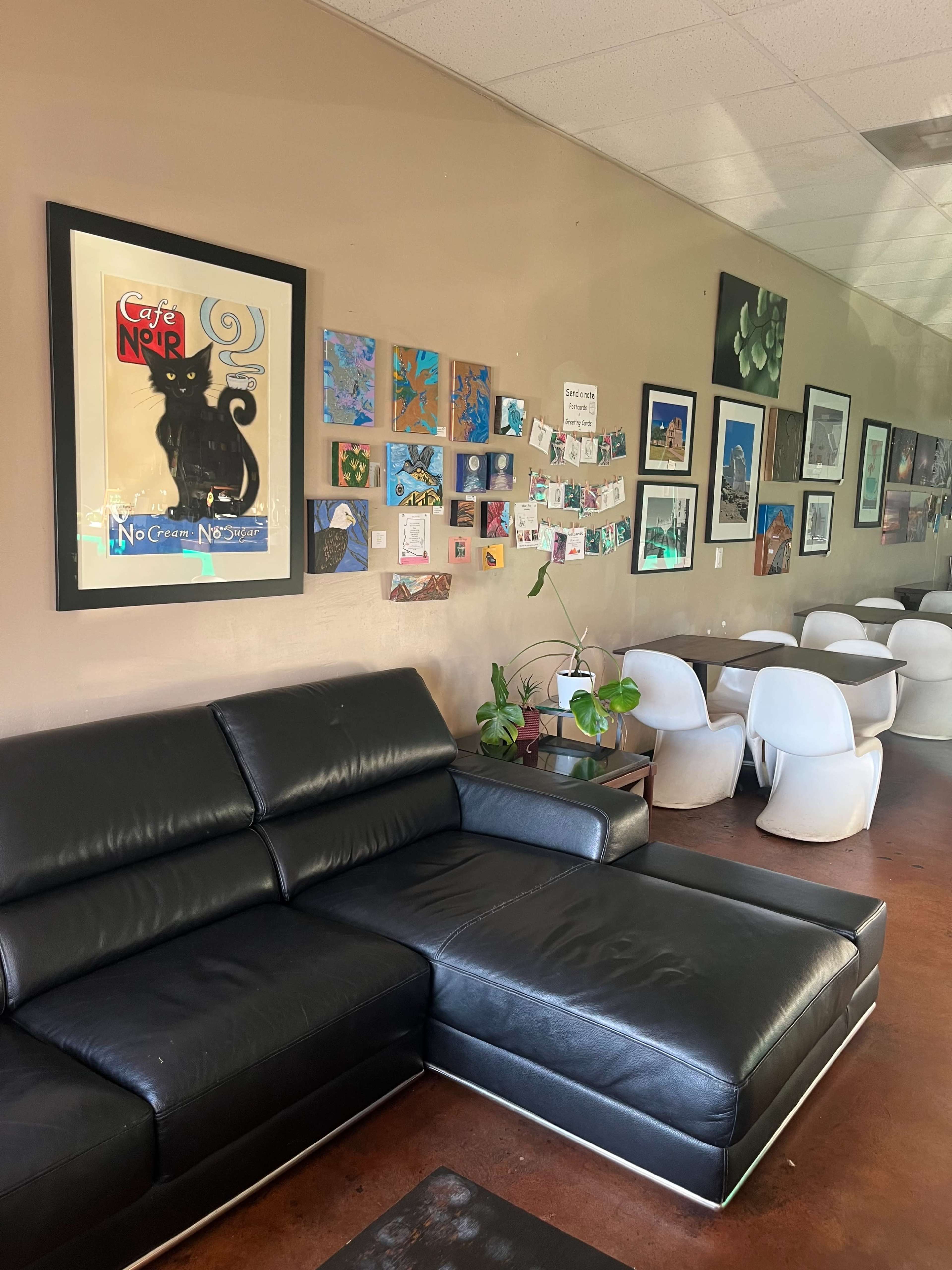 A cafe interior with a black leather sofa, a wall decorated with various framed artworks, and several tables and chairs in the background.
