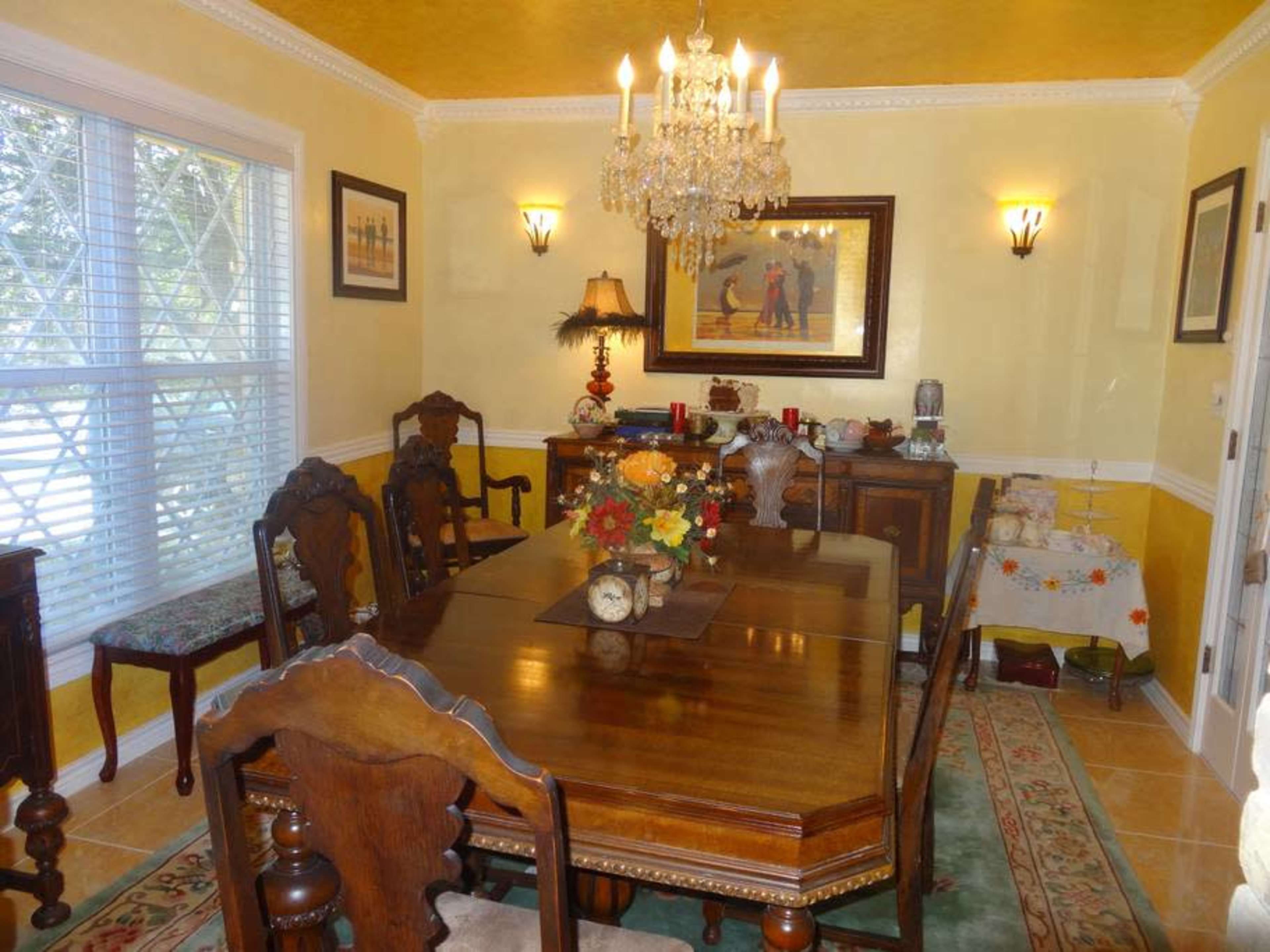 A dining room with a wooden table, eight carved chairs, a chandelier, and yellow walls adorned with artwork and decorative items.