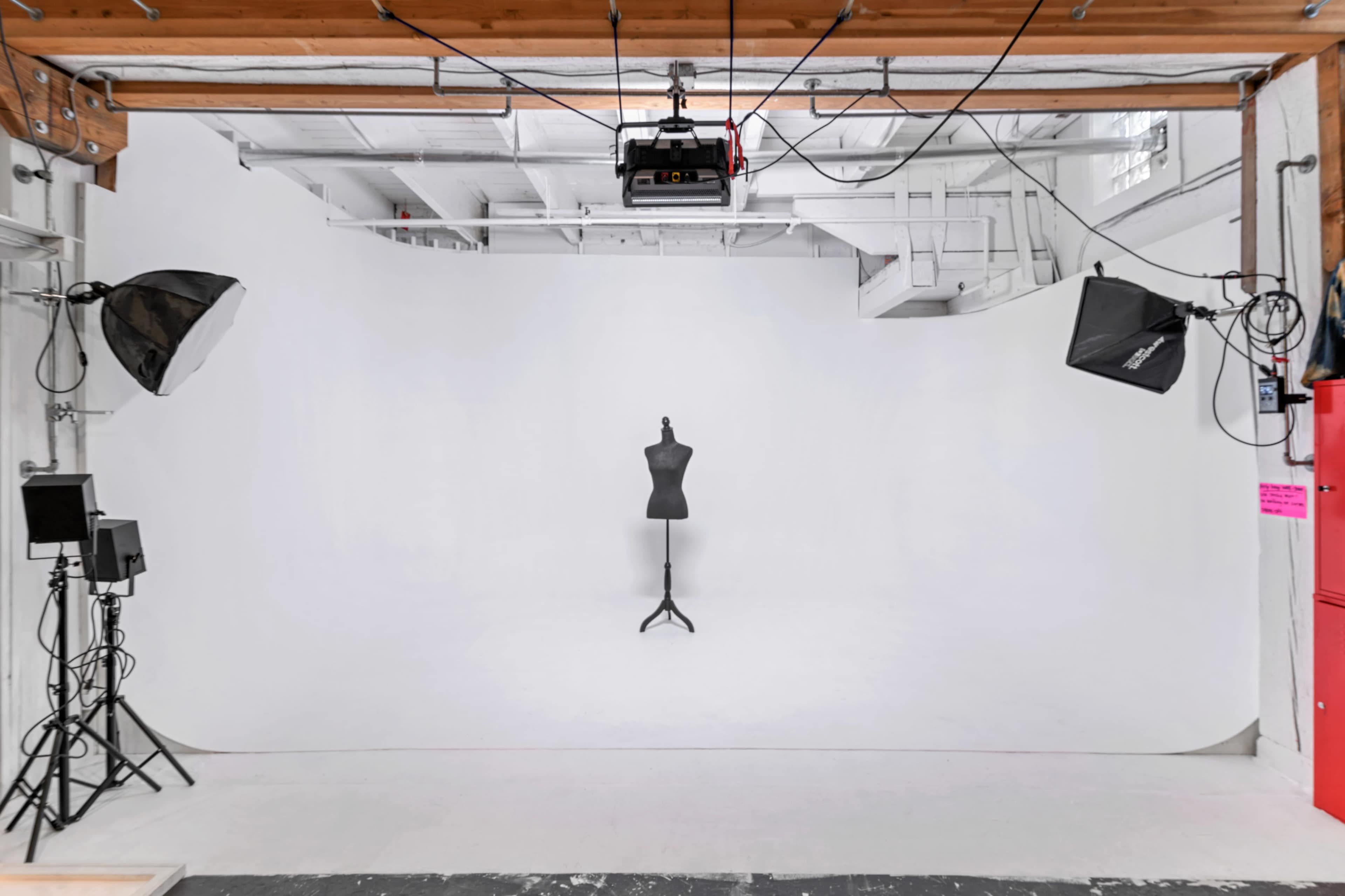 A photography studio features a white backdrop, a dress form on a stand in the center, and various lighting equipment arranged around the space.