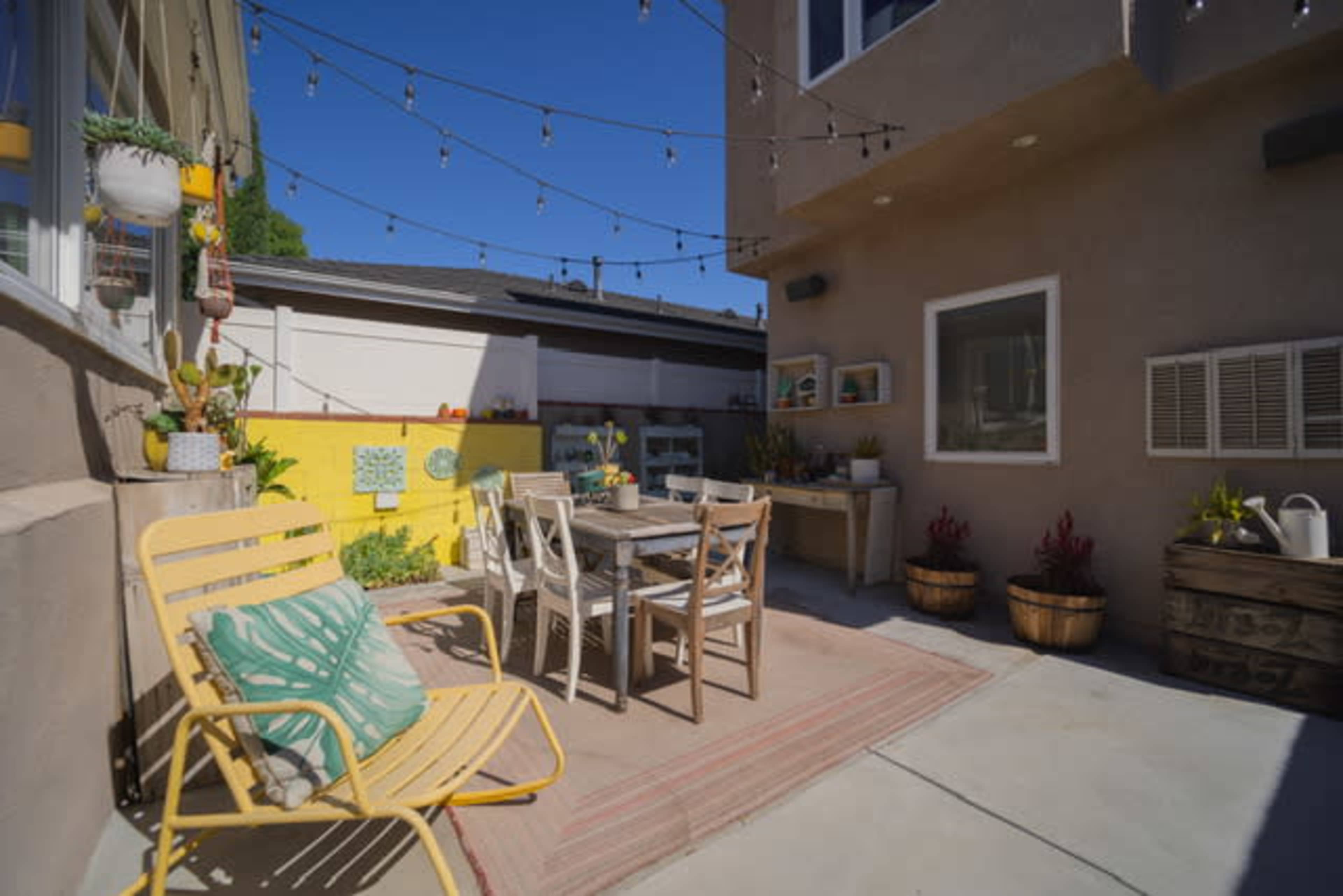 A small outdoor patio area features a table and chairs, surrounded by potted plants and string lights overhead.