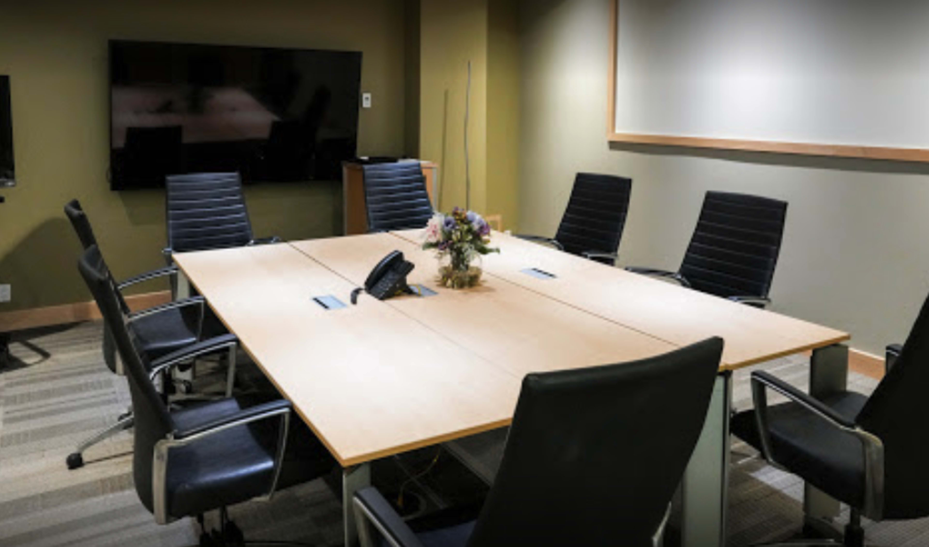 A conference room features a large wooden table surrounded by black chairs, with a television screen on the wall and a floral centerpiece.